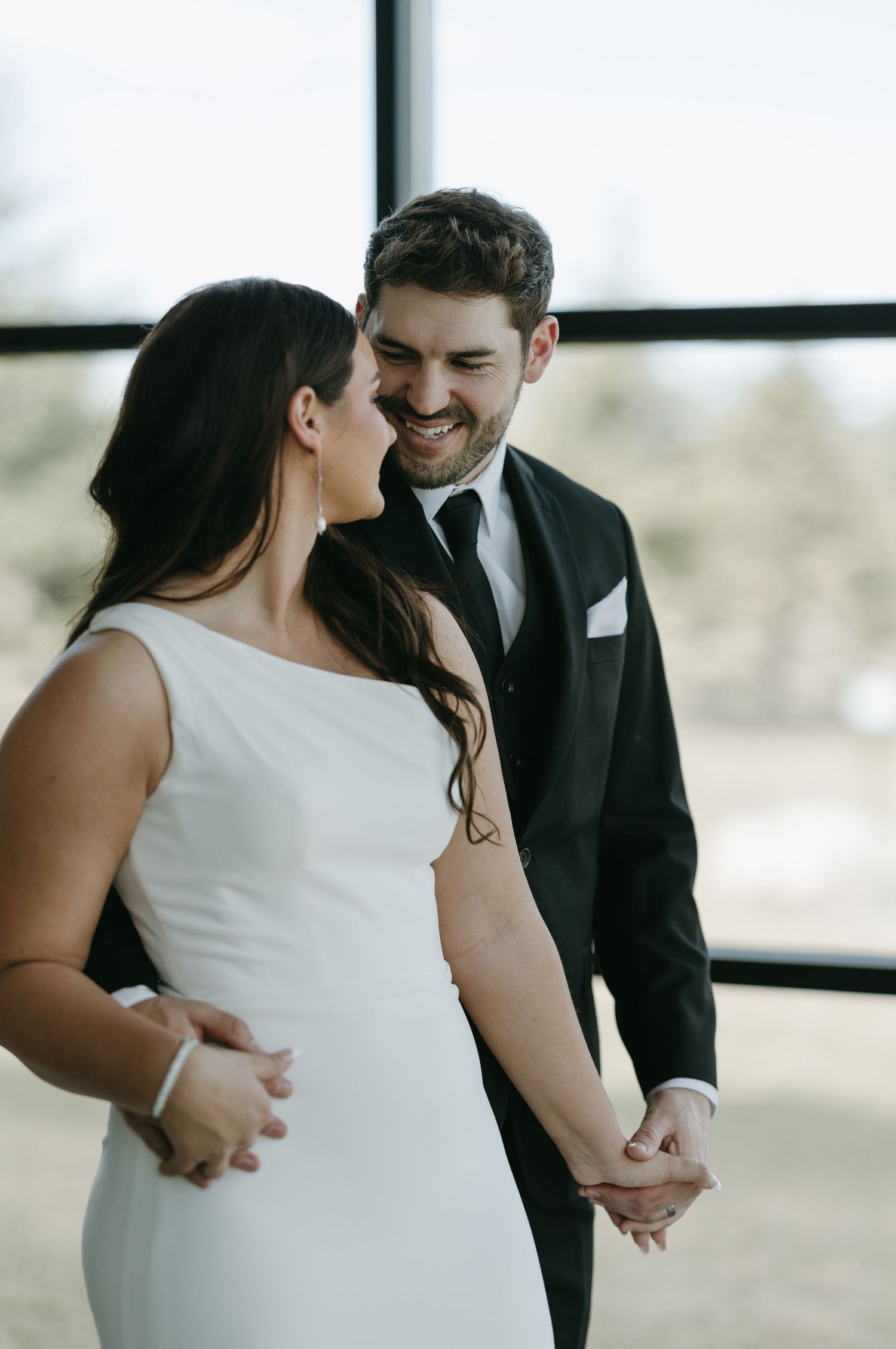 A candid moment between newlyweds sharing laughter near large windows in their wedding attire.