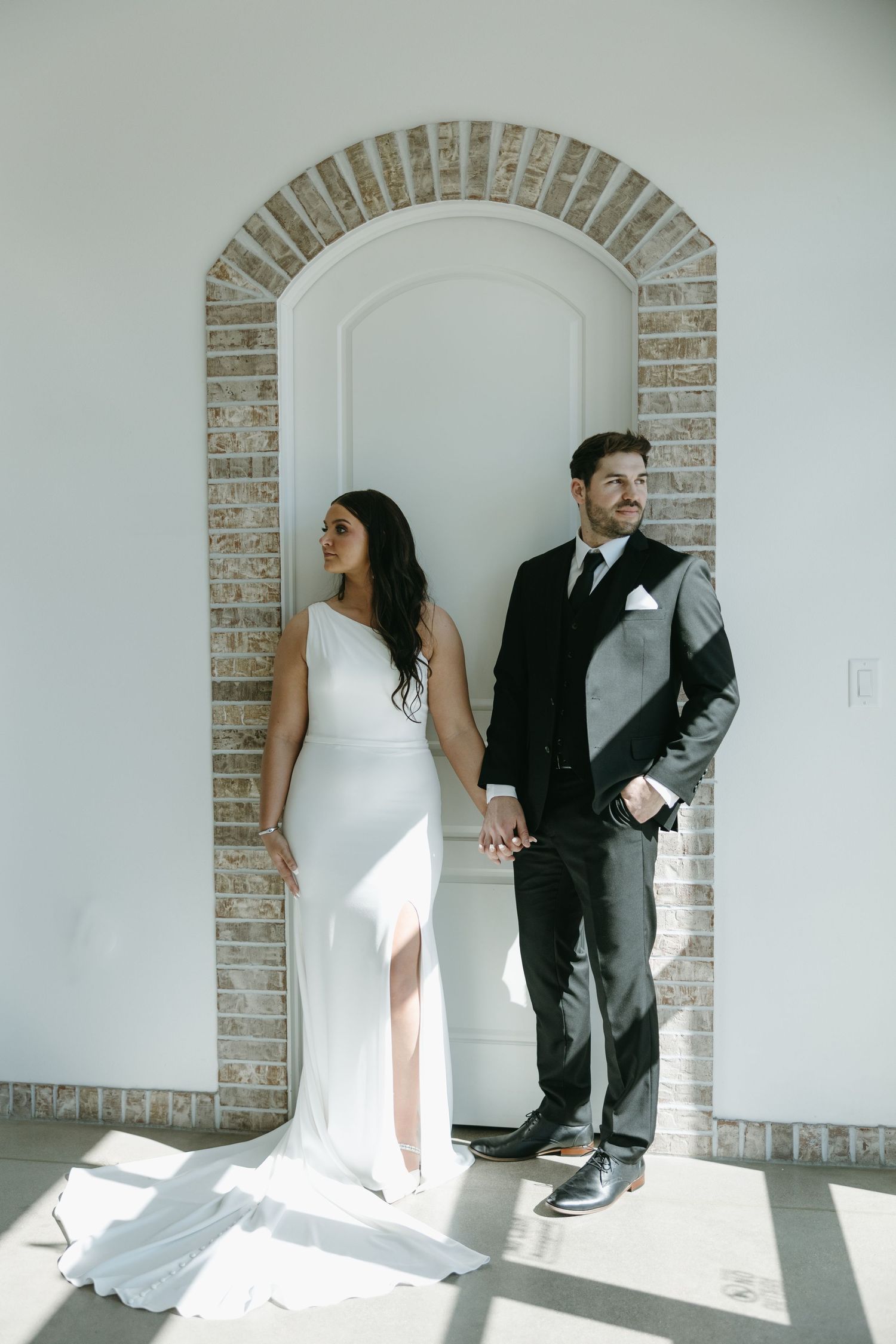 A stylish couple in wedding attire poses under a white brick archway with dramatic natural lighting.
