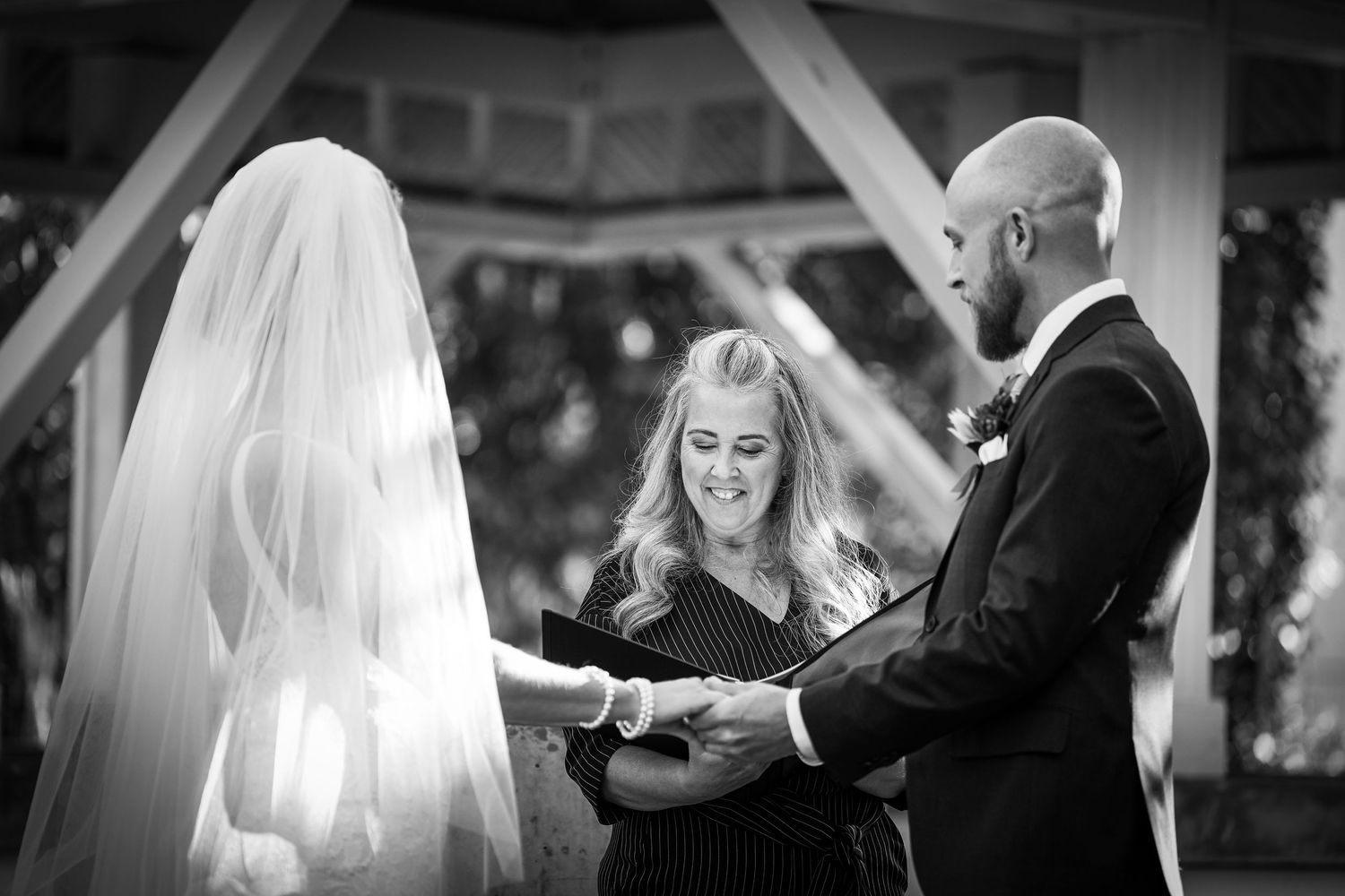 Black and white photo of a wedding ceremony with a couple exchanging vows in front of an officiant under an arch.