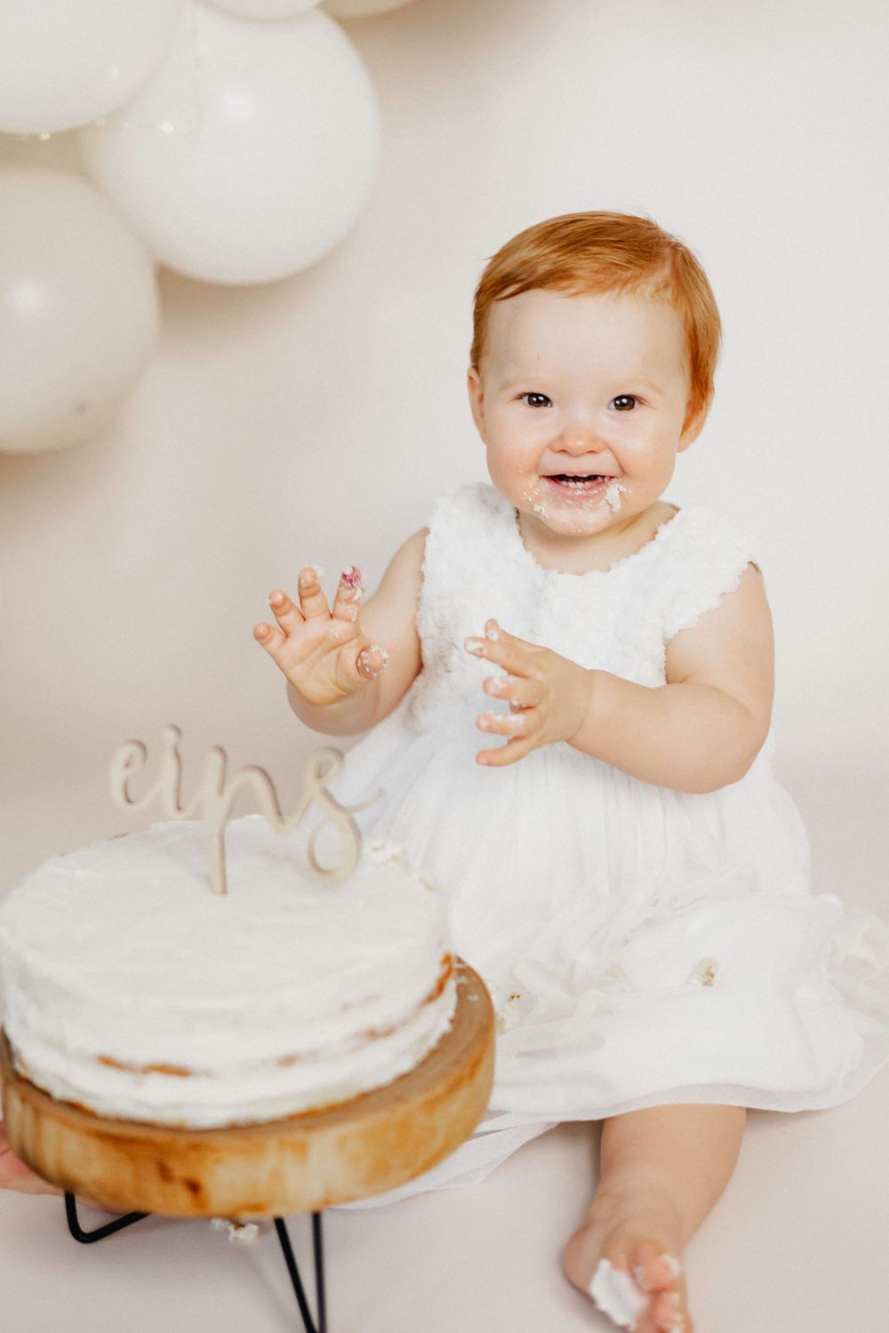 Ein Baby in einem weißen Kleid sitzt neben einer weißen Geburtstagstorte mit Luftballondekoration im Hintergrund.
