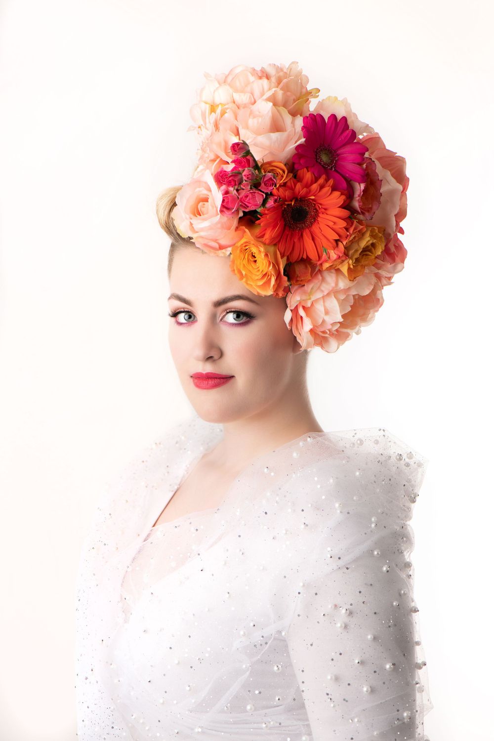A stunning floral headpiece with peach, orange and magenta blooms adorns a white lace outfit against light background.