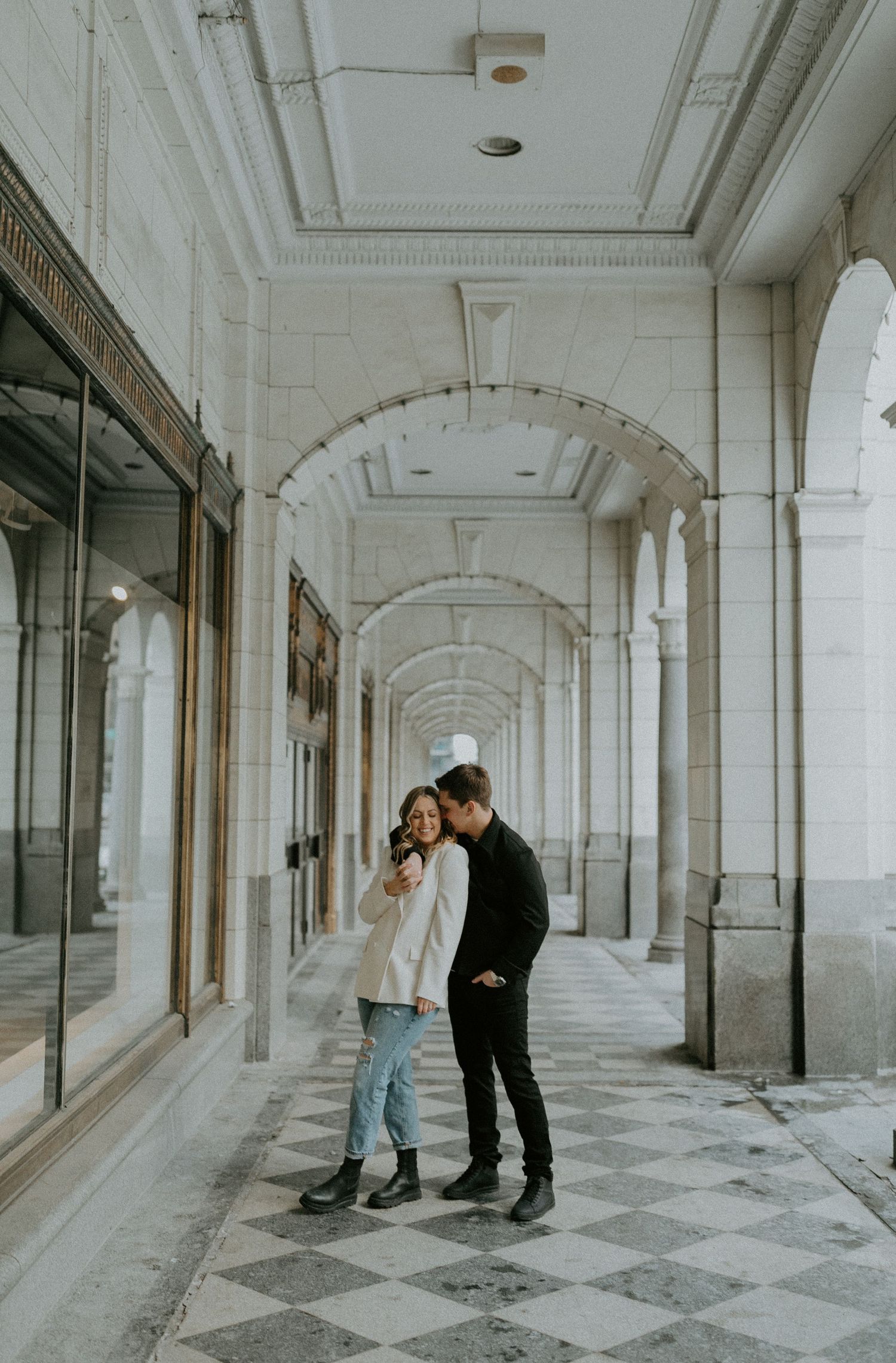 A couple shares an intimate moment beneath an elegant white arched colonnade with checkered floor tiles.
