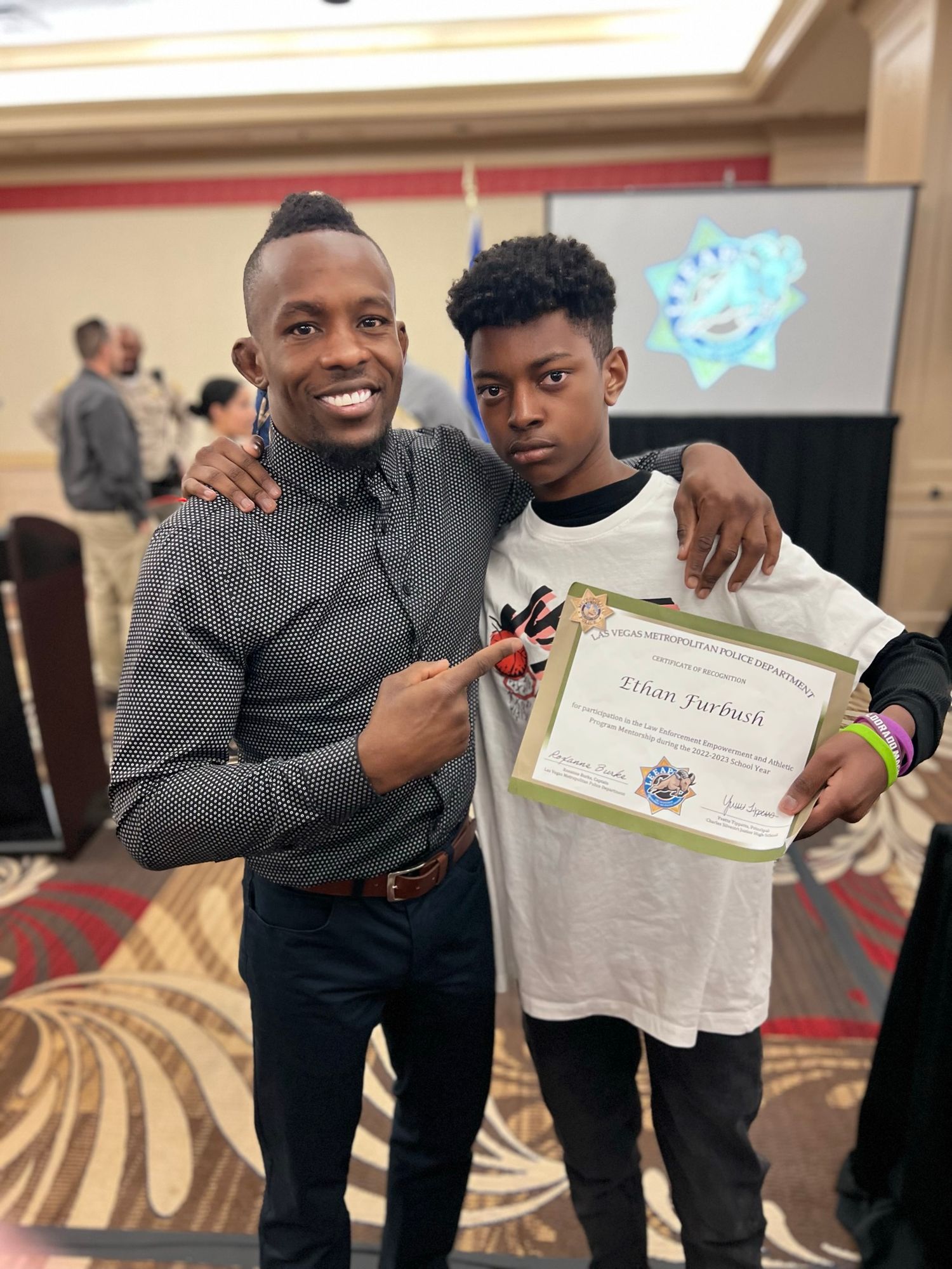 Two people pose together at an event, with one holding up a certificate while the other shows a small object.