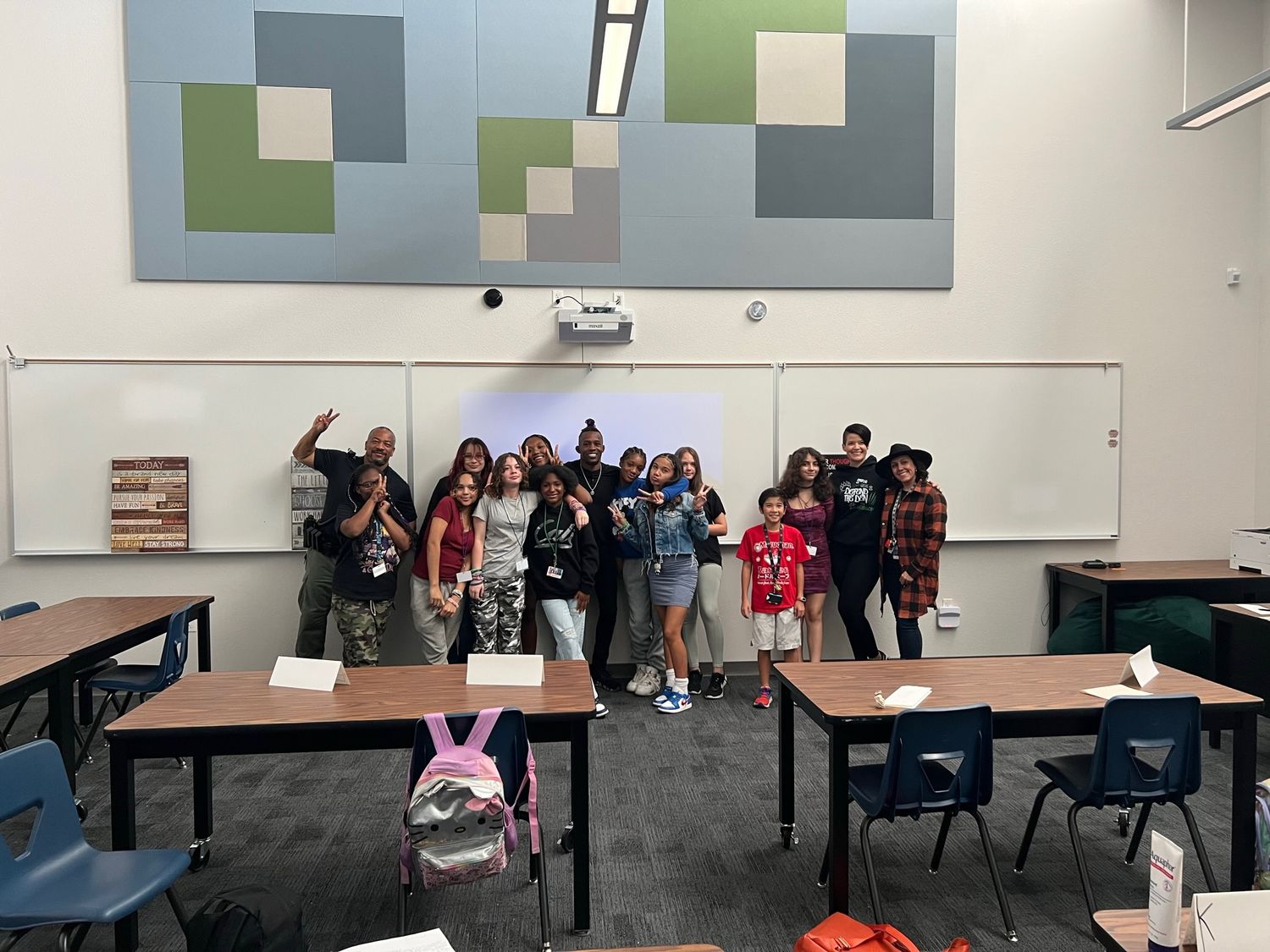 Students pose together in a classroom with desks and a geometric wall art display above a whiteboard.