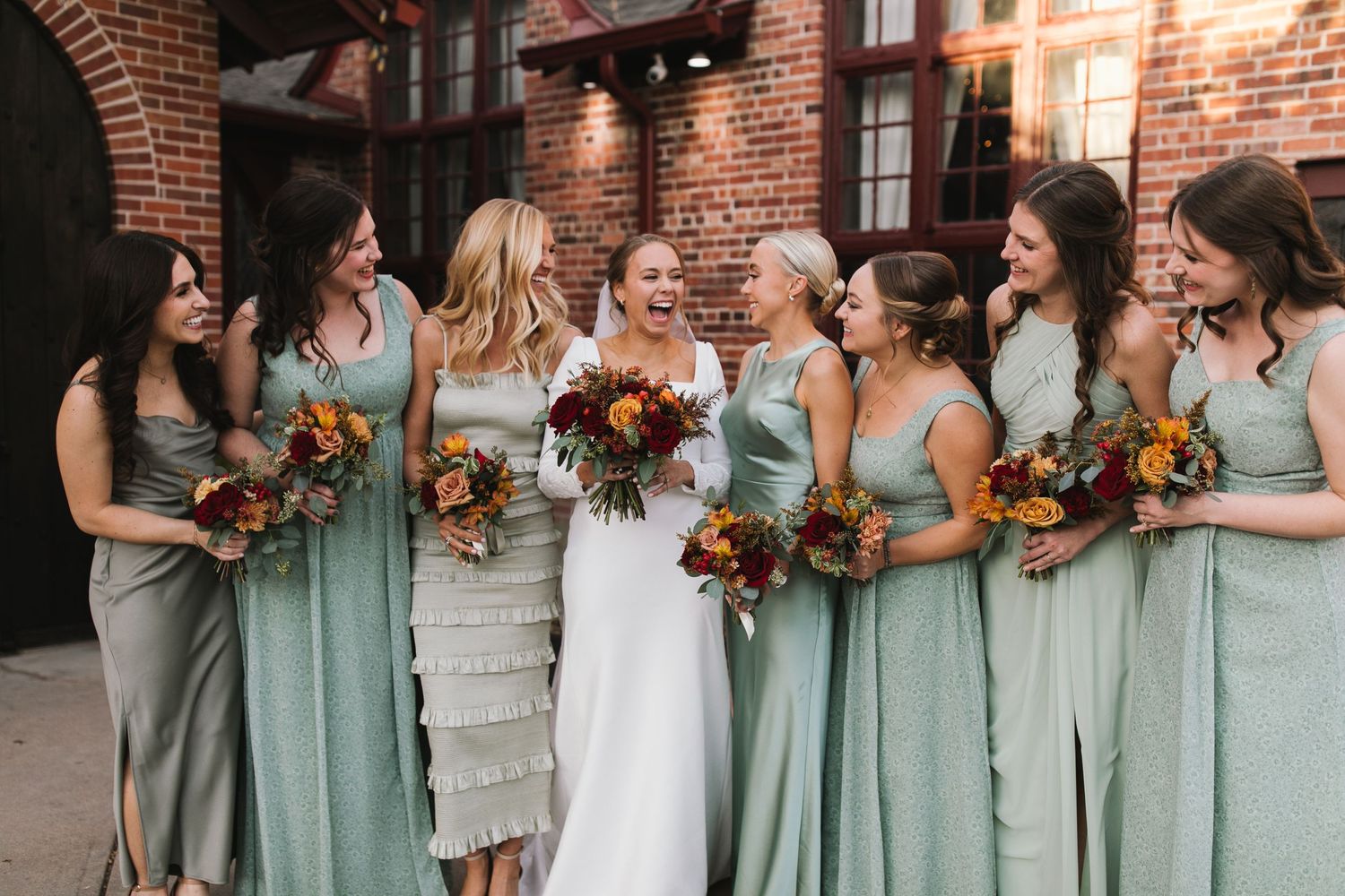 Wedding party in sage green bridesmaid dresses holding autumn-colored bouquets.