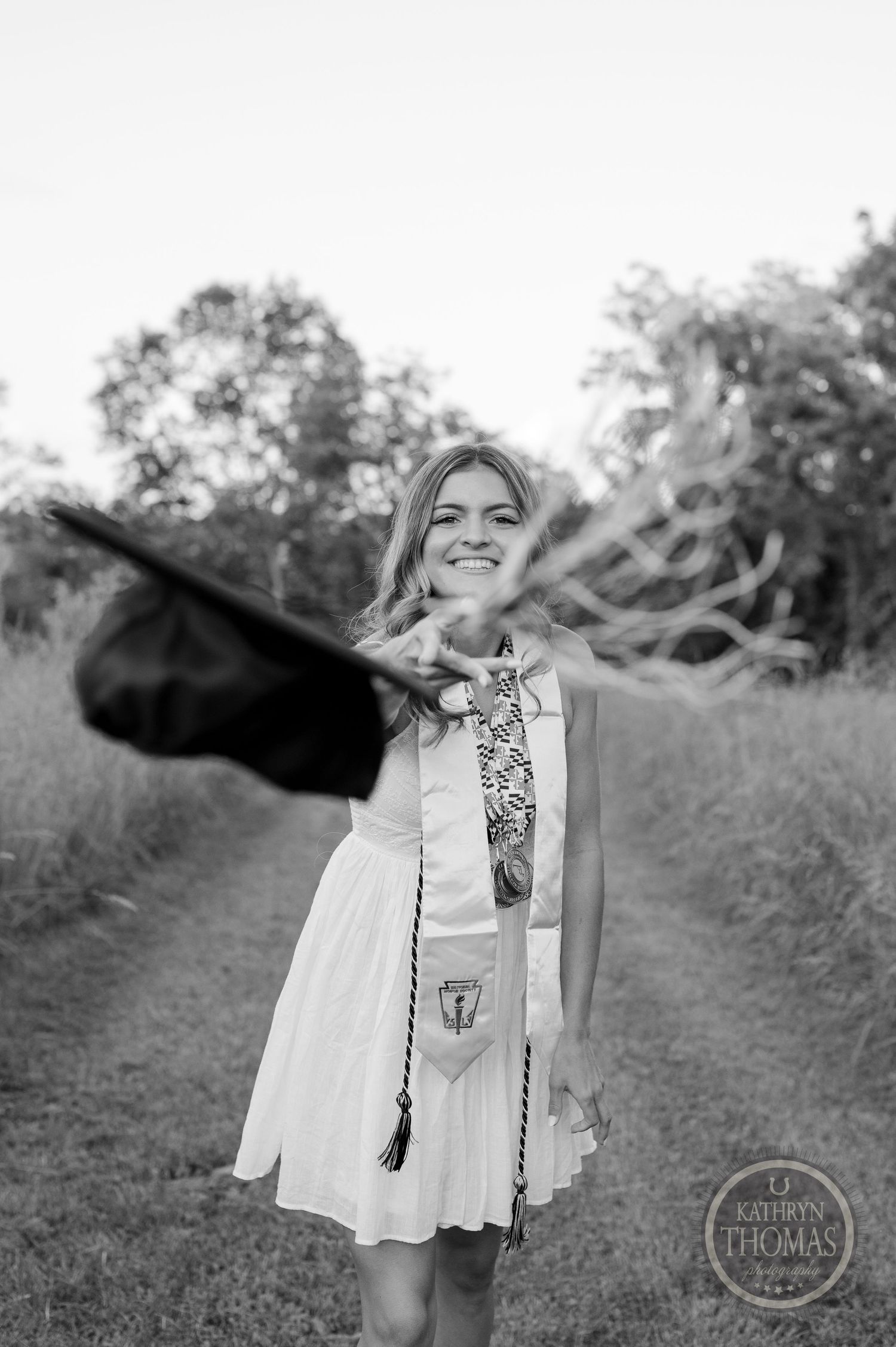 A graduation photoshoot in a field with a white sundress and black graduation cap being tossed in the air.