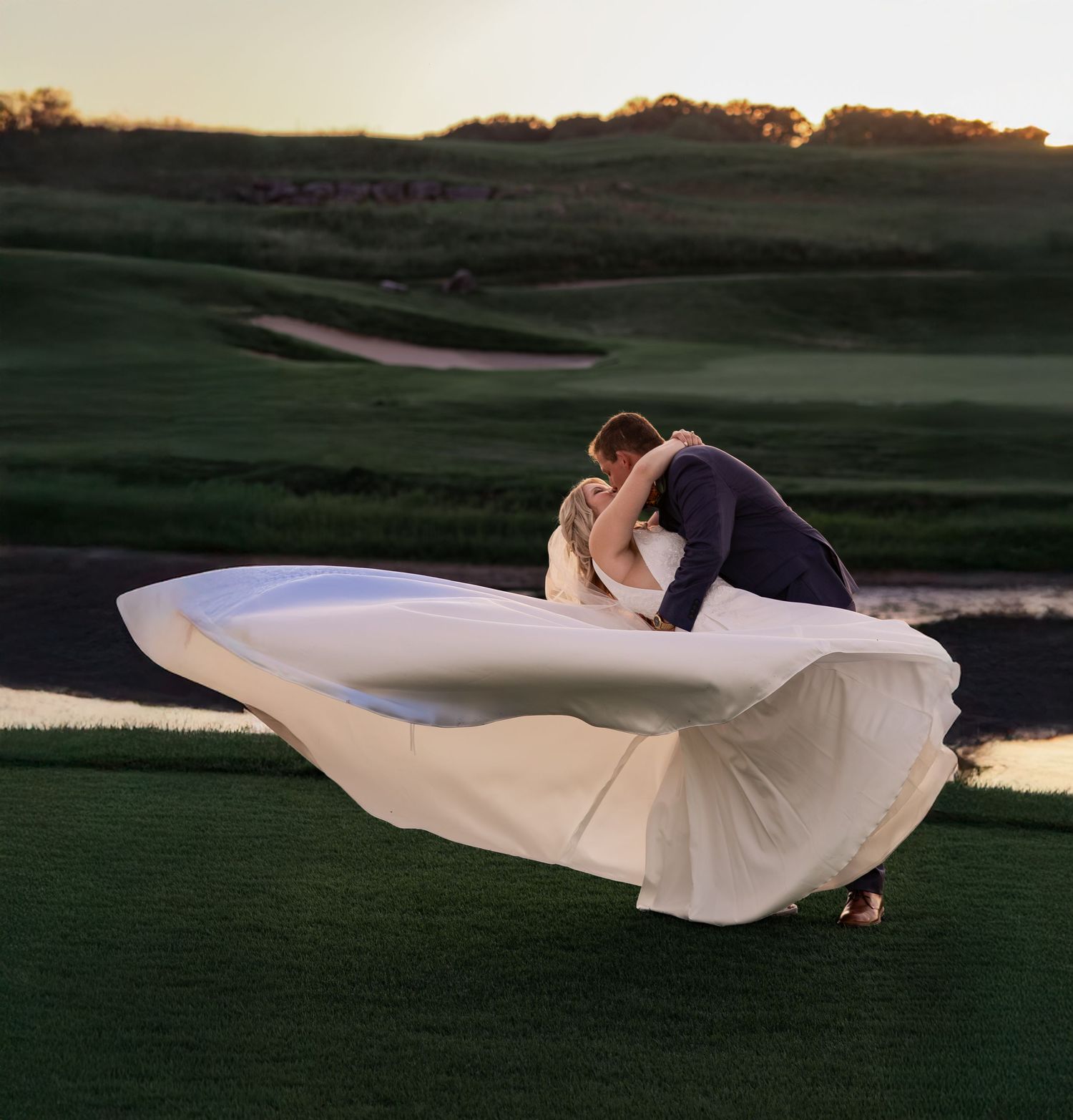 A flowing white fabric billows dramatically across a grassy field at sunset creating an ethereal scene.