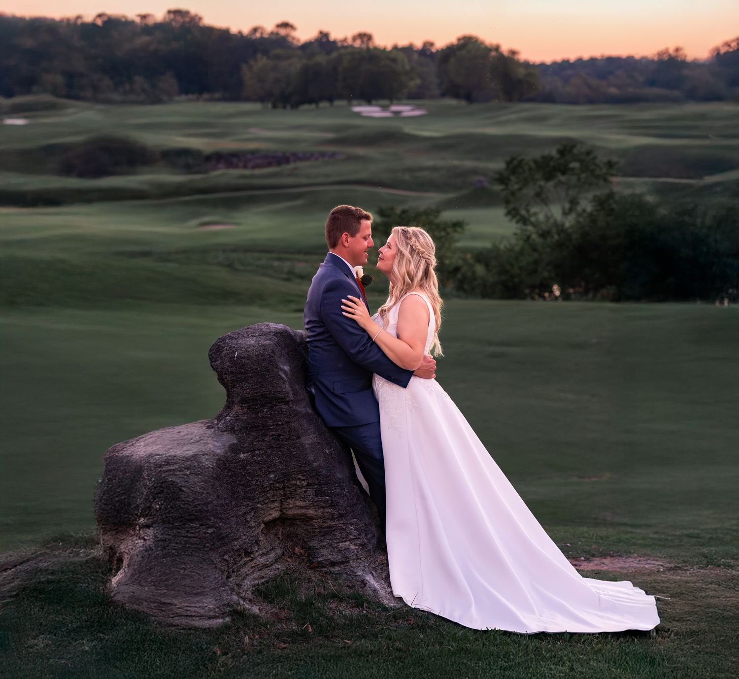 A couple in wedding attire embrace on a large rock overlooking a scenic golf course at sunset.