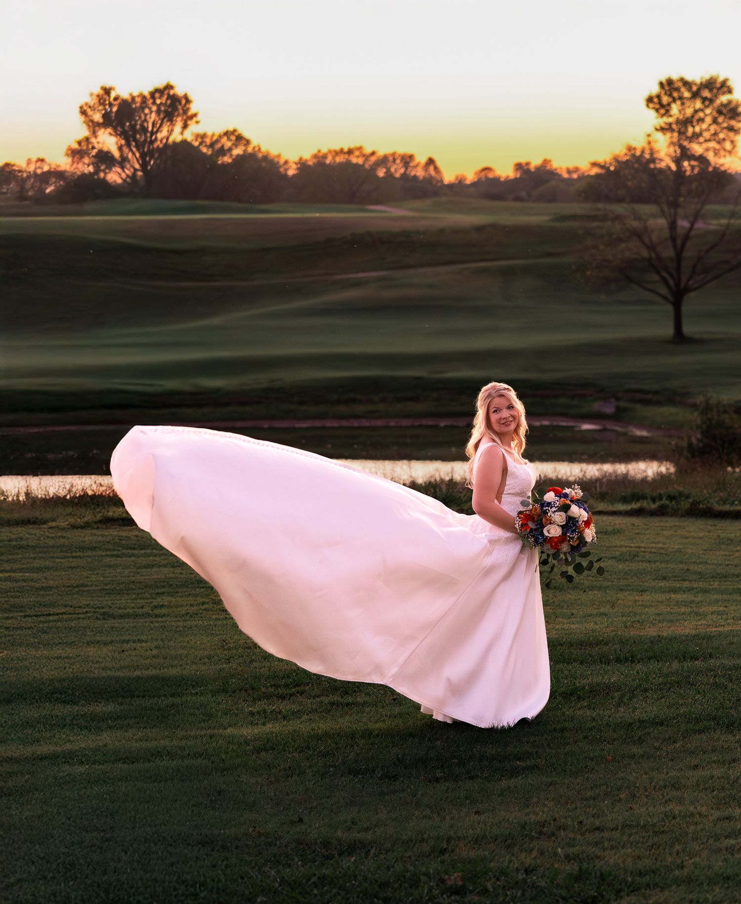 A person in a flowing white wedding dress stands against a sunset backdrop on rolling hills with their veil floating in the wind.