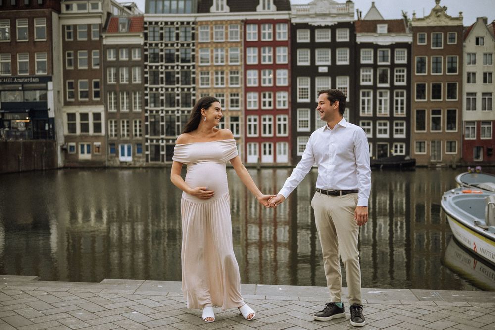 Pregnant couple holding hands in front of colorful houses and canal in Amsterdam