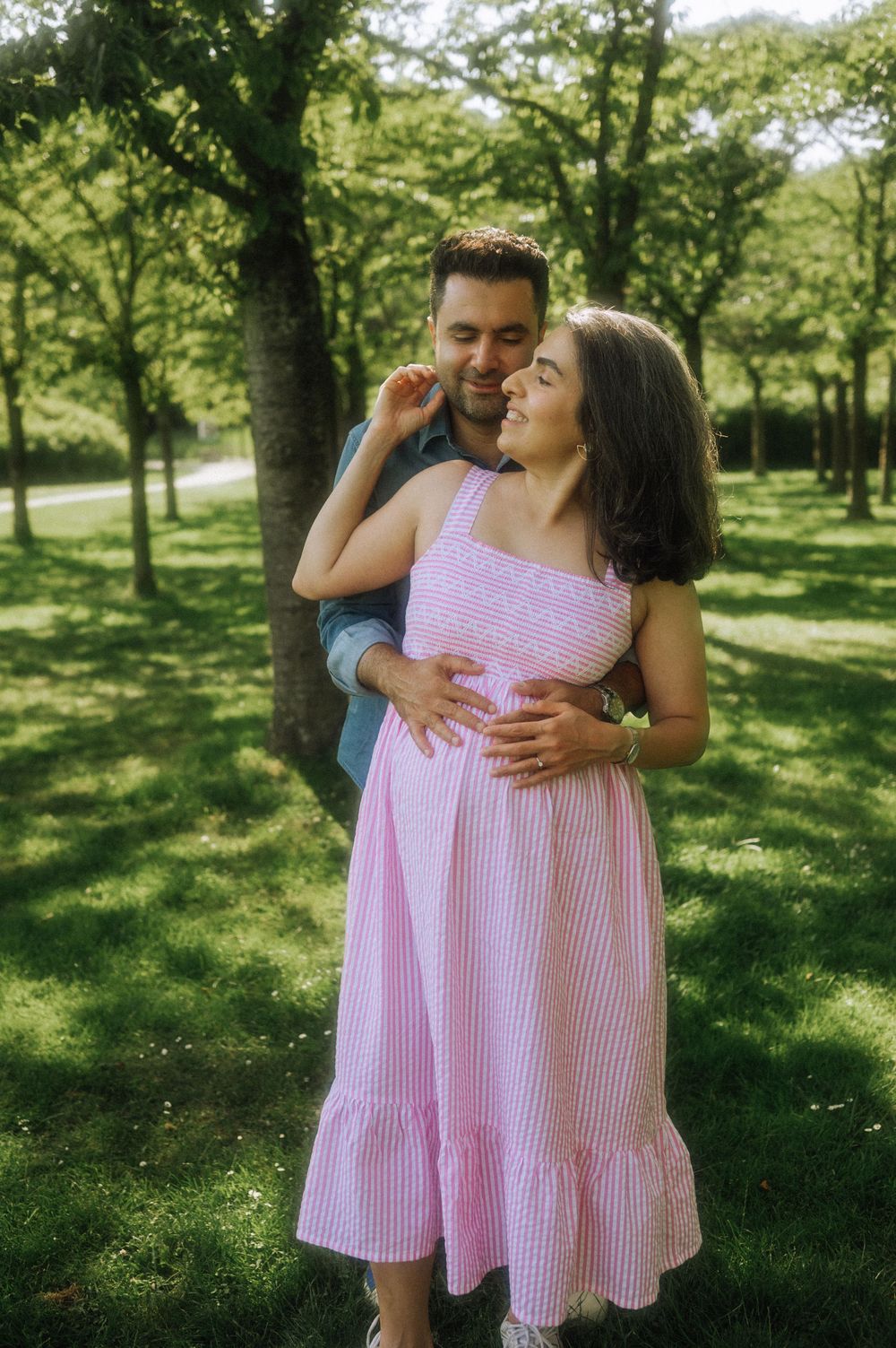 A couple embraces in a sunny park setting with lush green trees in the background.