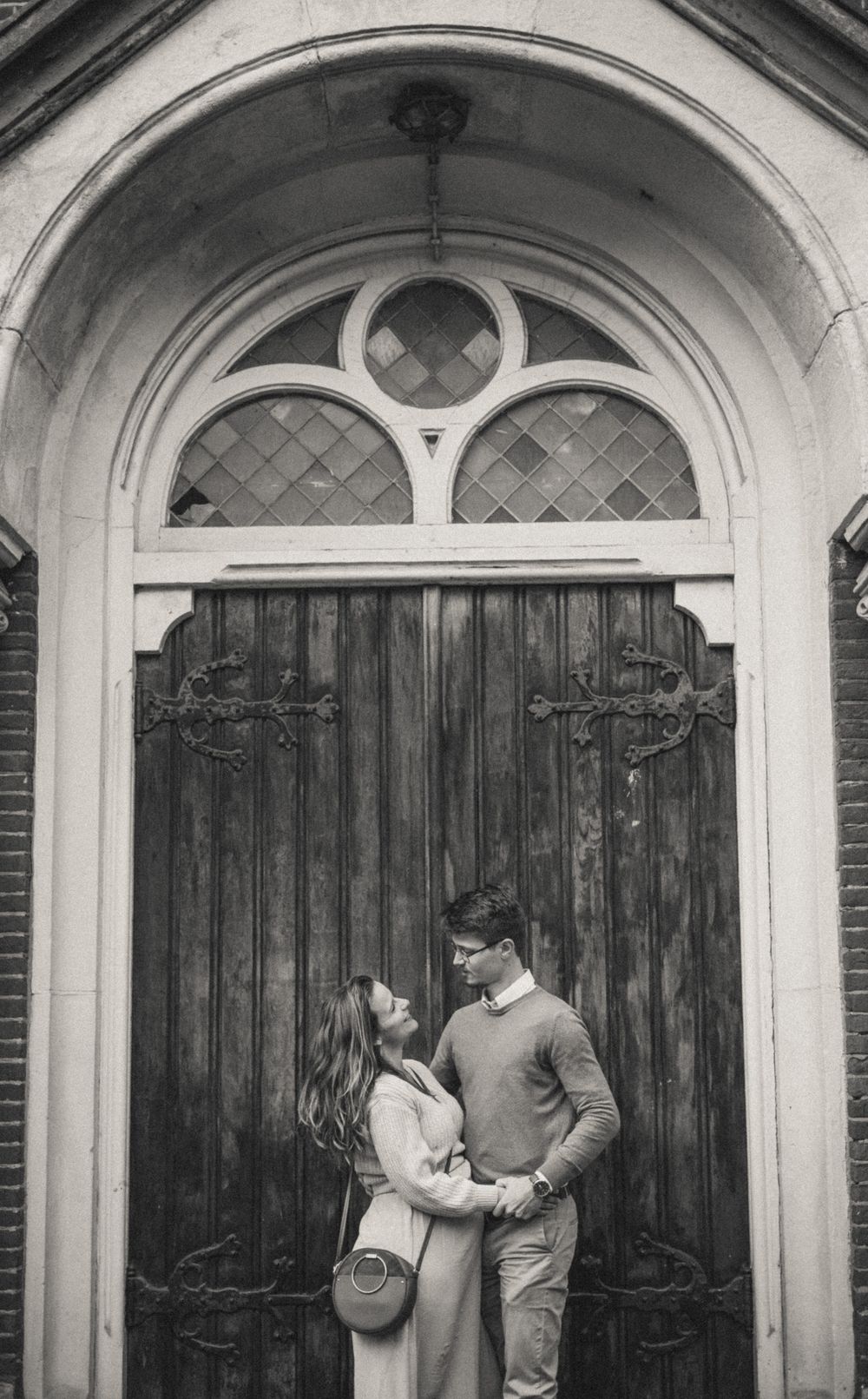 Romantic couple embracing in front of ornate wooden church doors with arched windows.