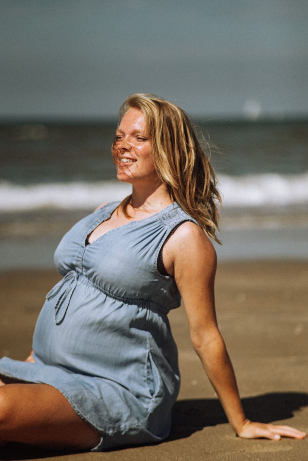Woman in gray dress relaxing on sandy beach near ocean waves.