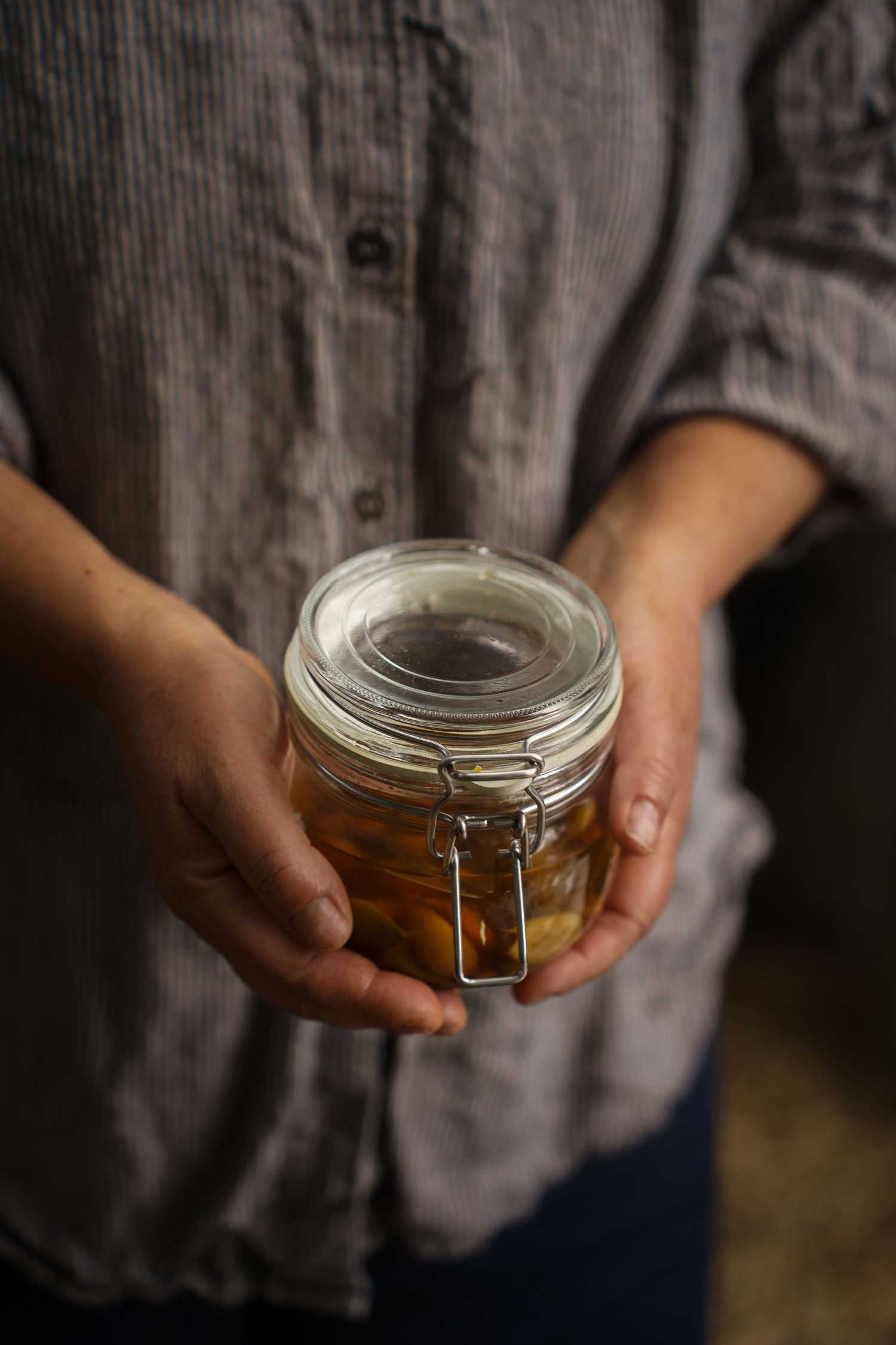 Person in a striped linen shirt holding a glass jar filled with preserved amber liquid.