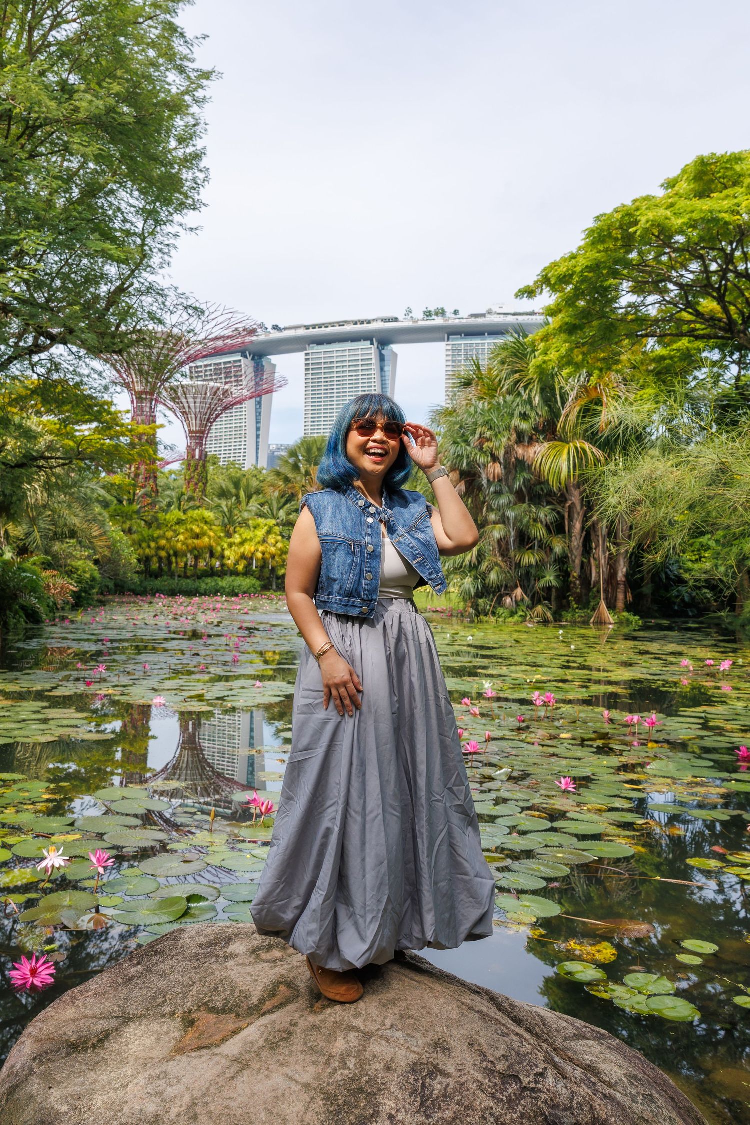 A person poses in a denim vest and flowy grey skirt at Gardens by the Bay Singapore with lotus pond and Marina Bay Sands visible.
