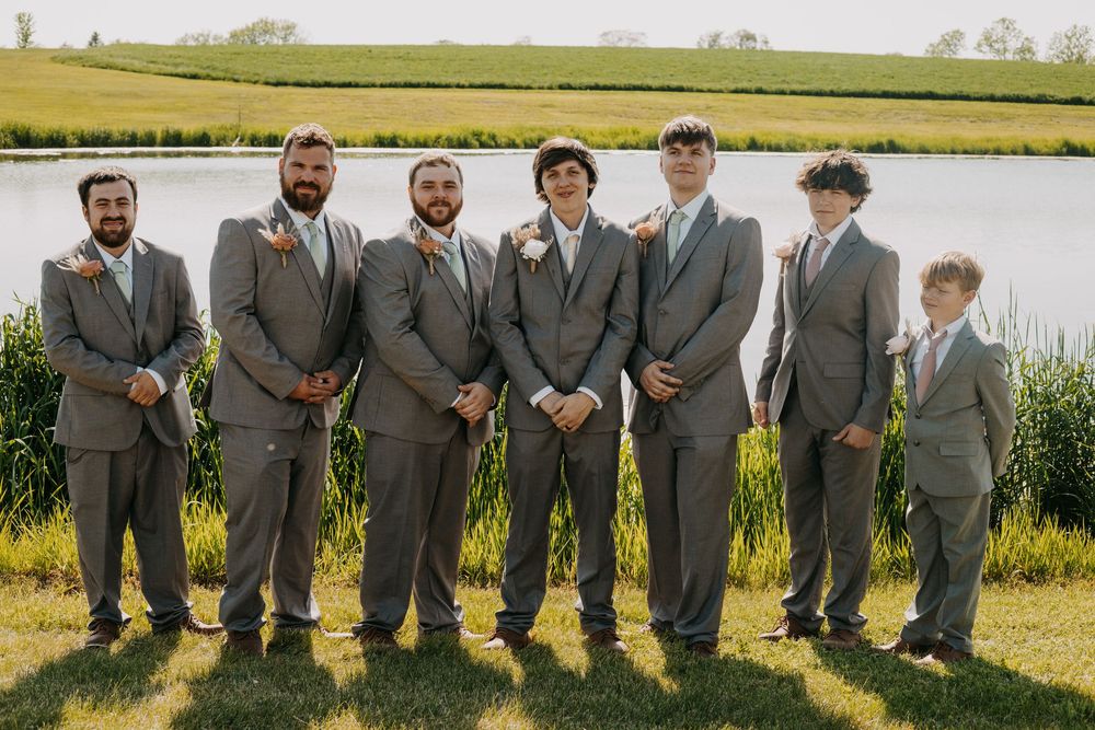 A row of people wearing matching gray suits stand together outdoors in a grassy field on a sunny day.