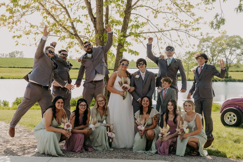 Wedding party poses playfully for group photo outdoors by a lake with trees and greenery in the background.