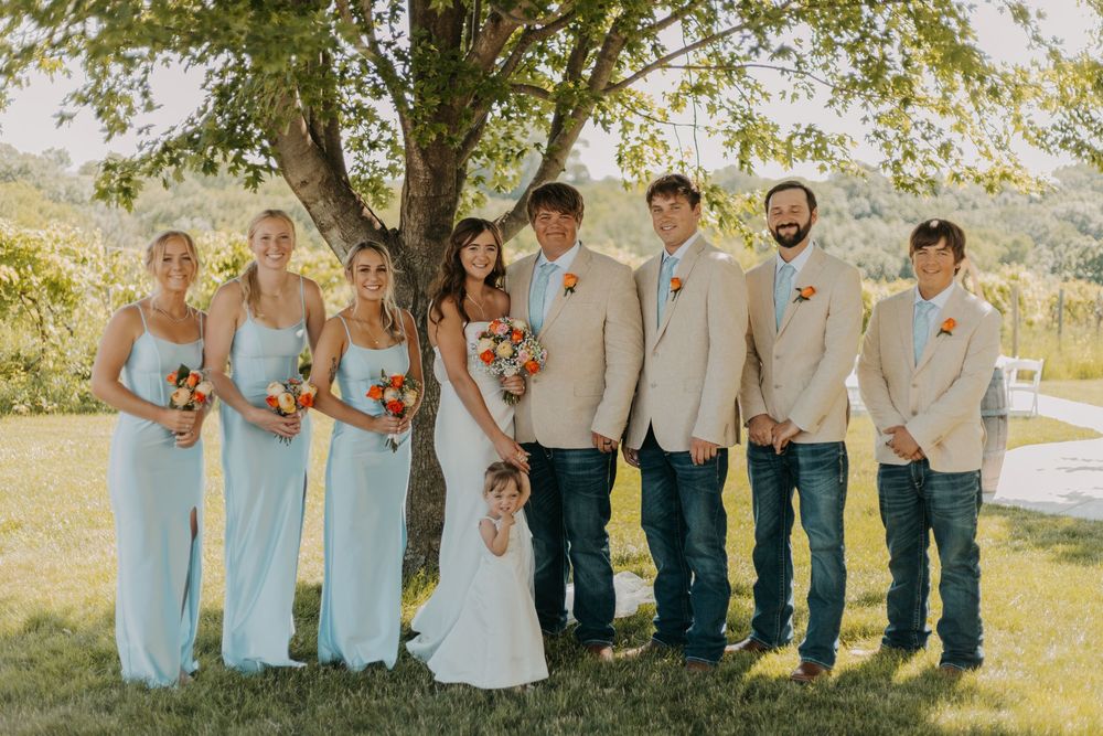 Wedding party poses outdoors in light blue and beige attire under trees at a rustic countryside venue.