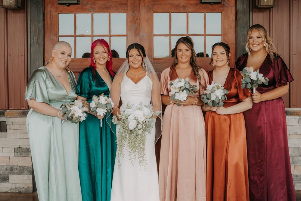 A wedding party poses in front of a rustic building wearing coordinated pastel and jewel-toned dresses.