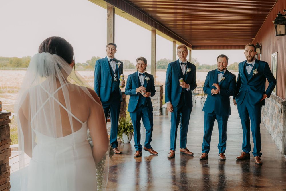 Wedding party in matching navy suits poses with drinks while bride watches from a covered porch.