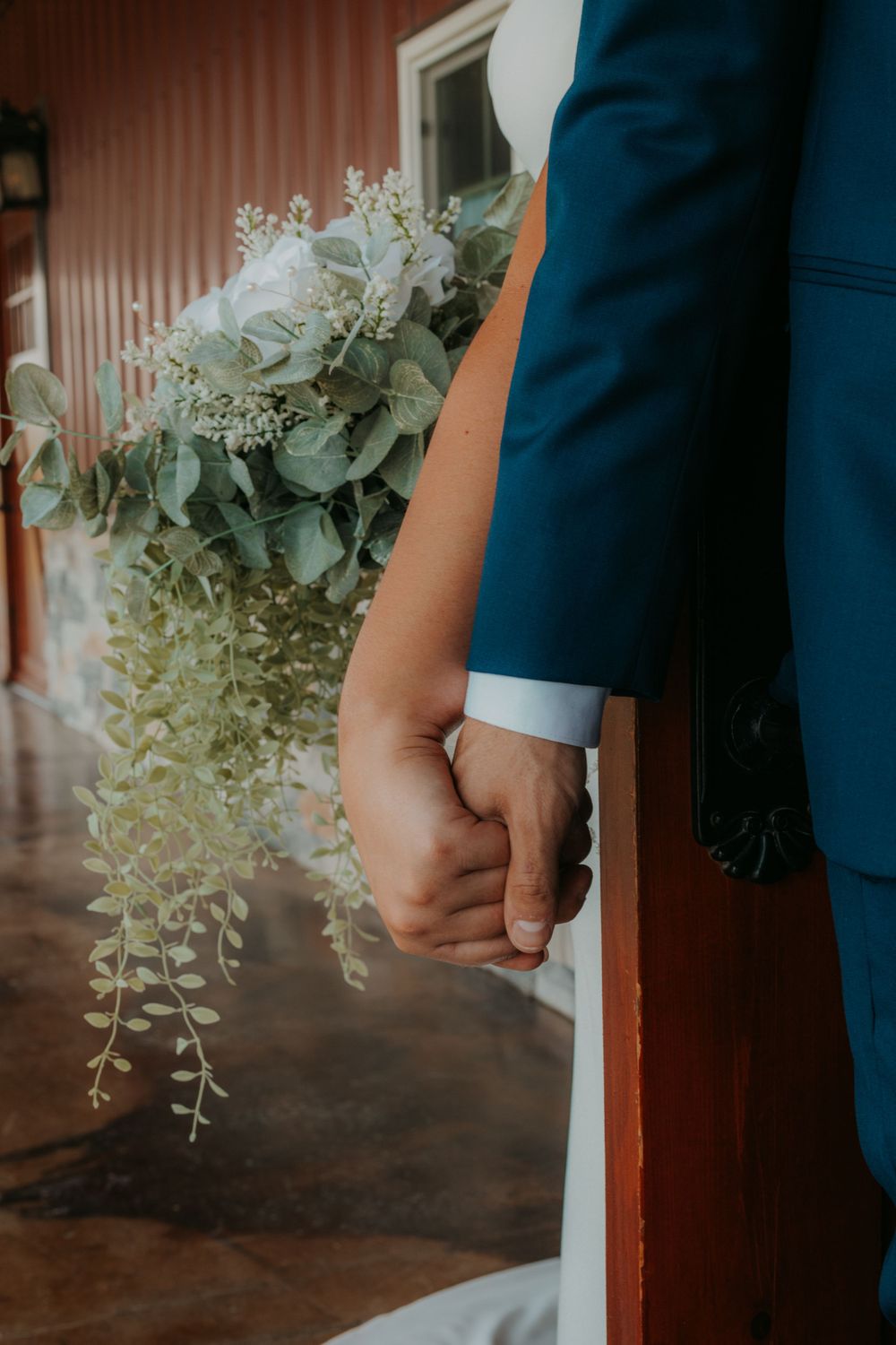 Close-up of newlyweds holding hands with cascading bridal bouquet against navy suit.