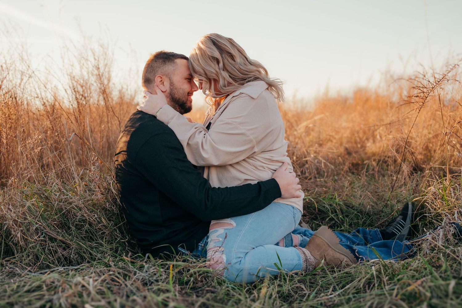 A couple embraces intimately while sitting in golden autumn grass during sunset.