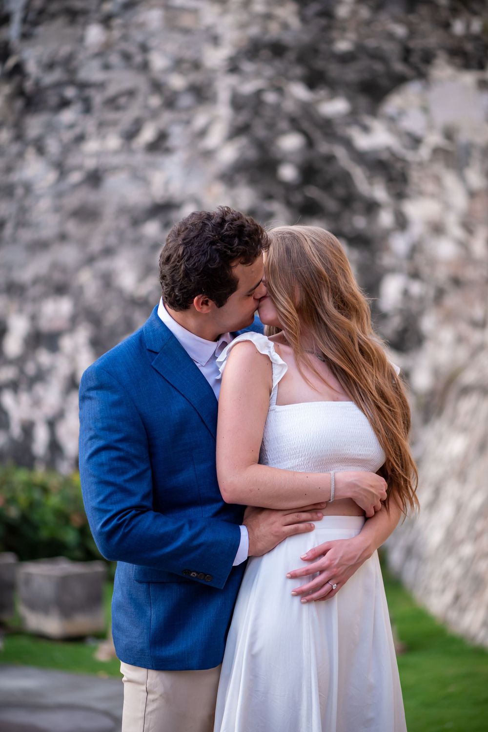 A couple embraces and kisses during an outdoor photoshoot in elegant formal attire.