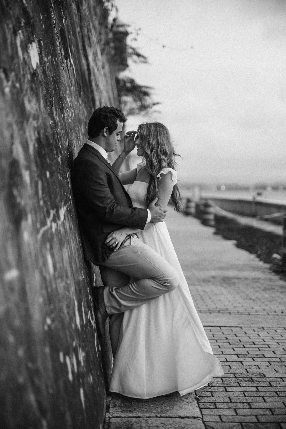 A black and white romantic moment captured against a stone wall by the waterfront.