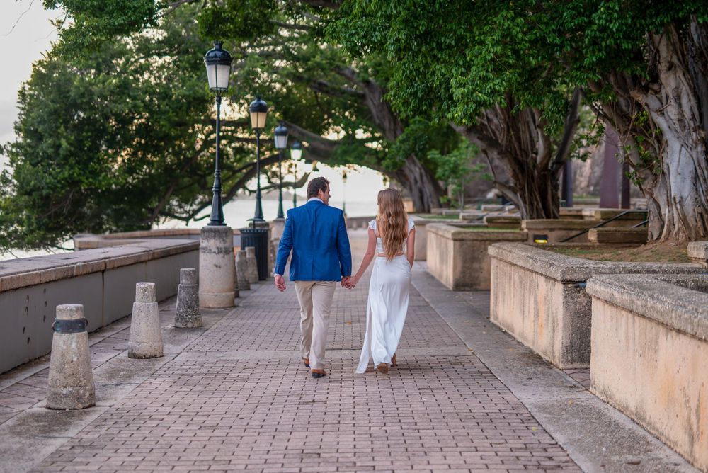 A couple walks hand in hand down a scenic stone pathway lined with trees and lampposts.