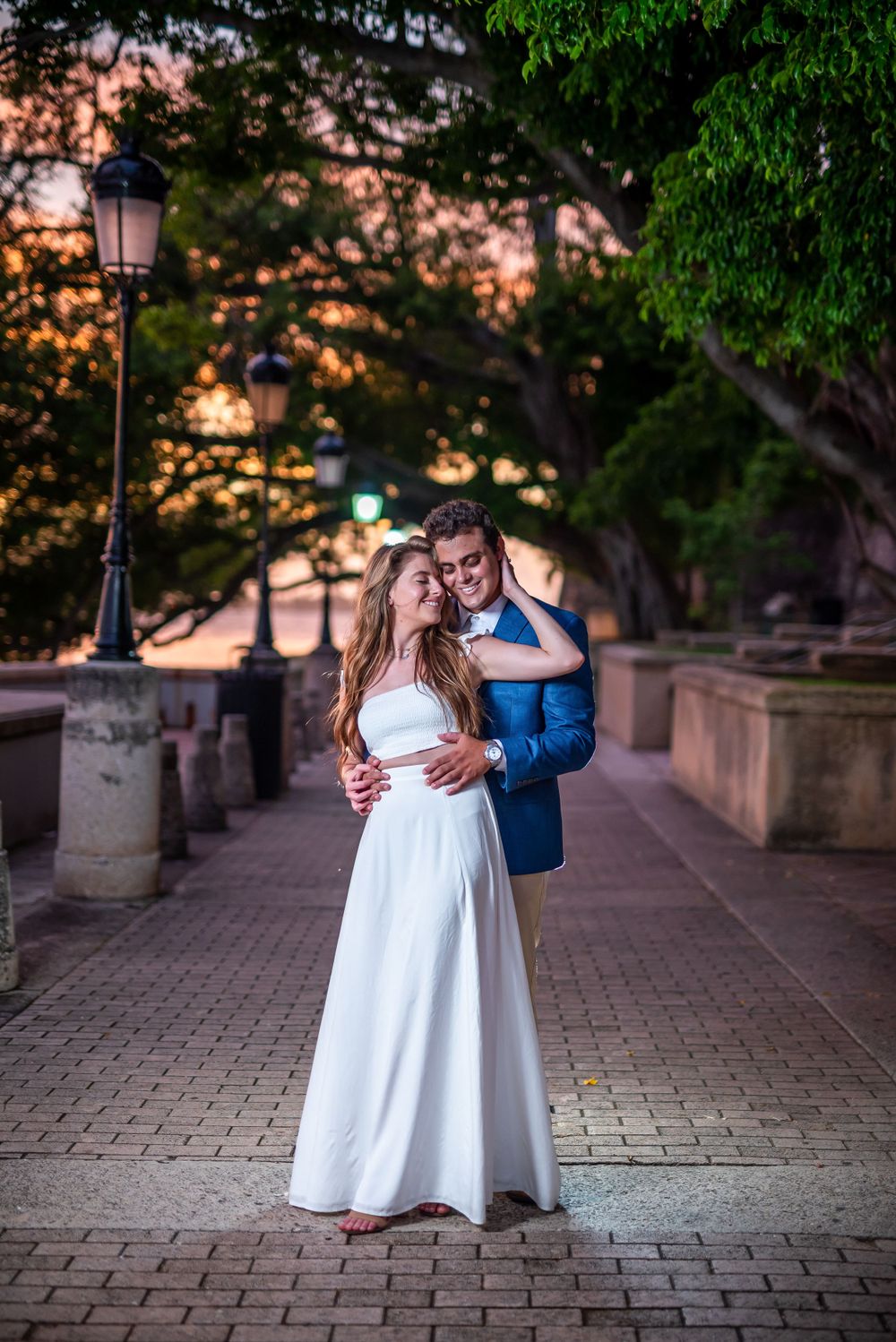 A sunset embrace on a charming brick pathway surrounded by trees and vintage lampposts.