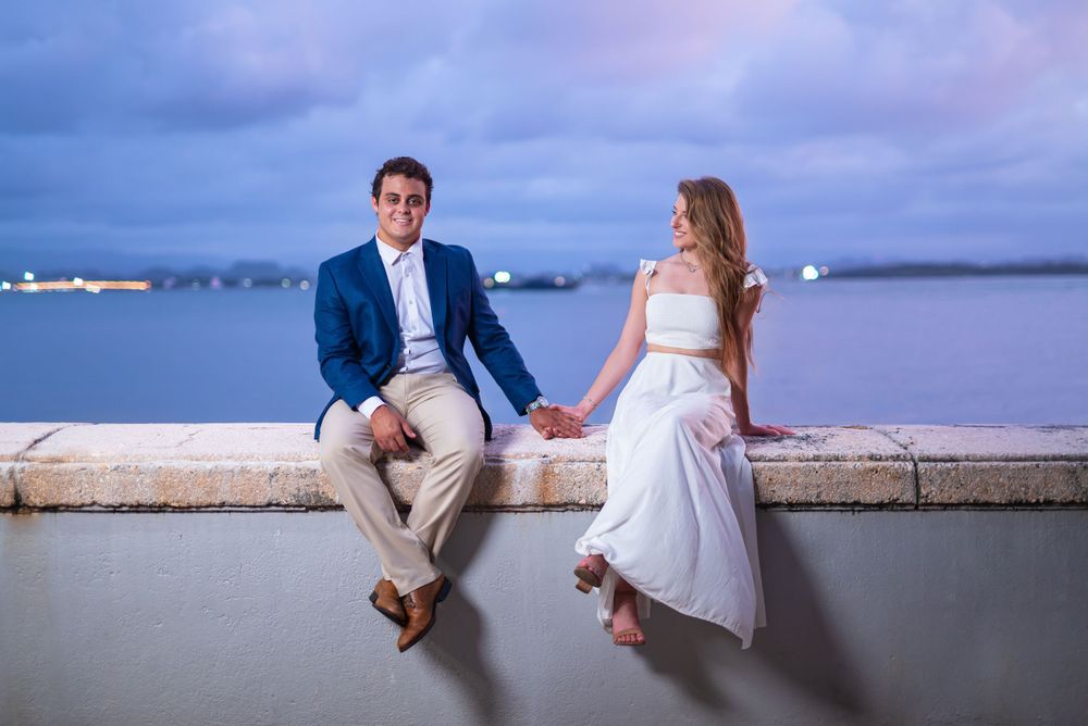 A couple sits together on a waterfront wall during blue hour with a harbor view.