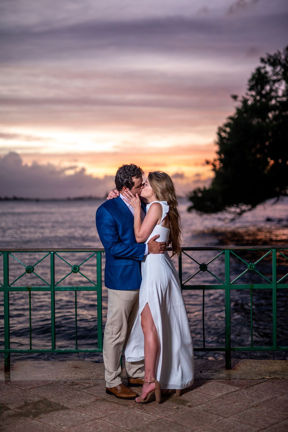 A couple embraces on a waterfront terrace at sunset with pink and purple skies over the ocean.