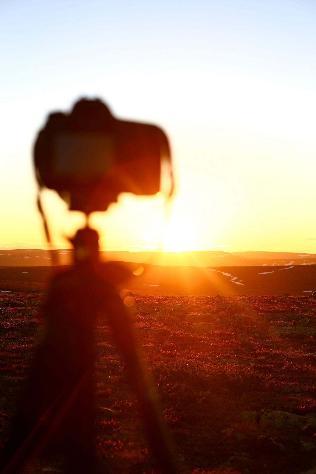 A silhouetted camera on a tripod captures a stunning golden sunset over a vast open landscape.