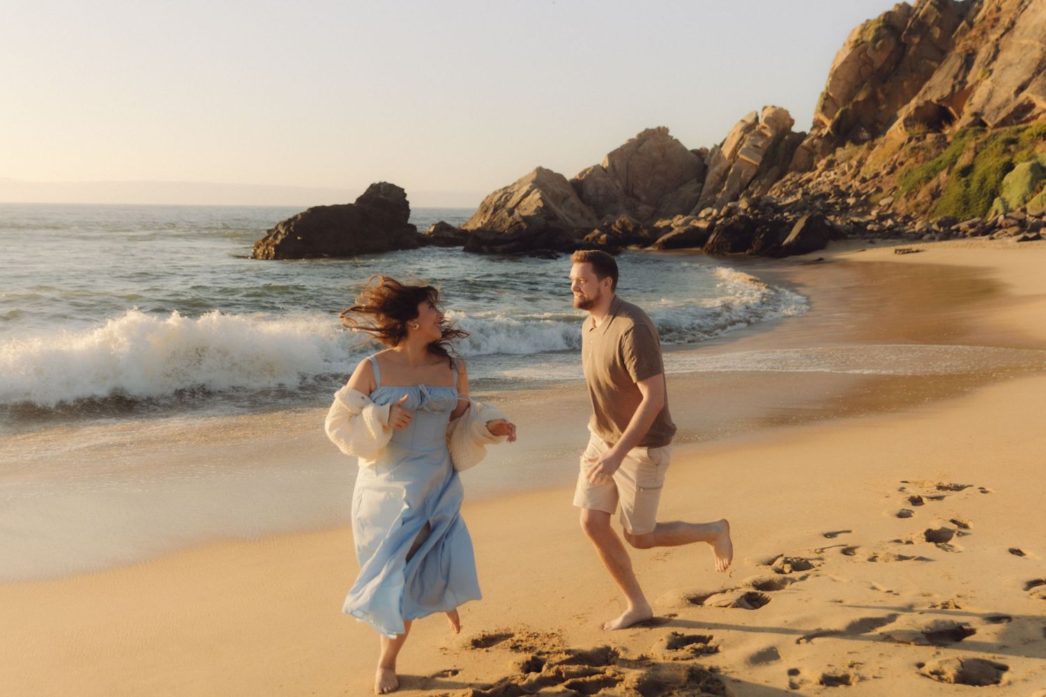 Couple running and laughing along a scenic rocky beach at golden hour sunset.