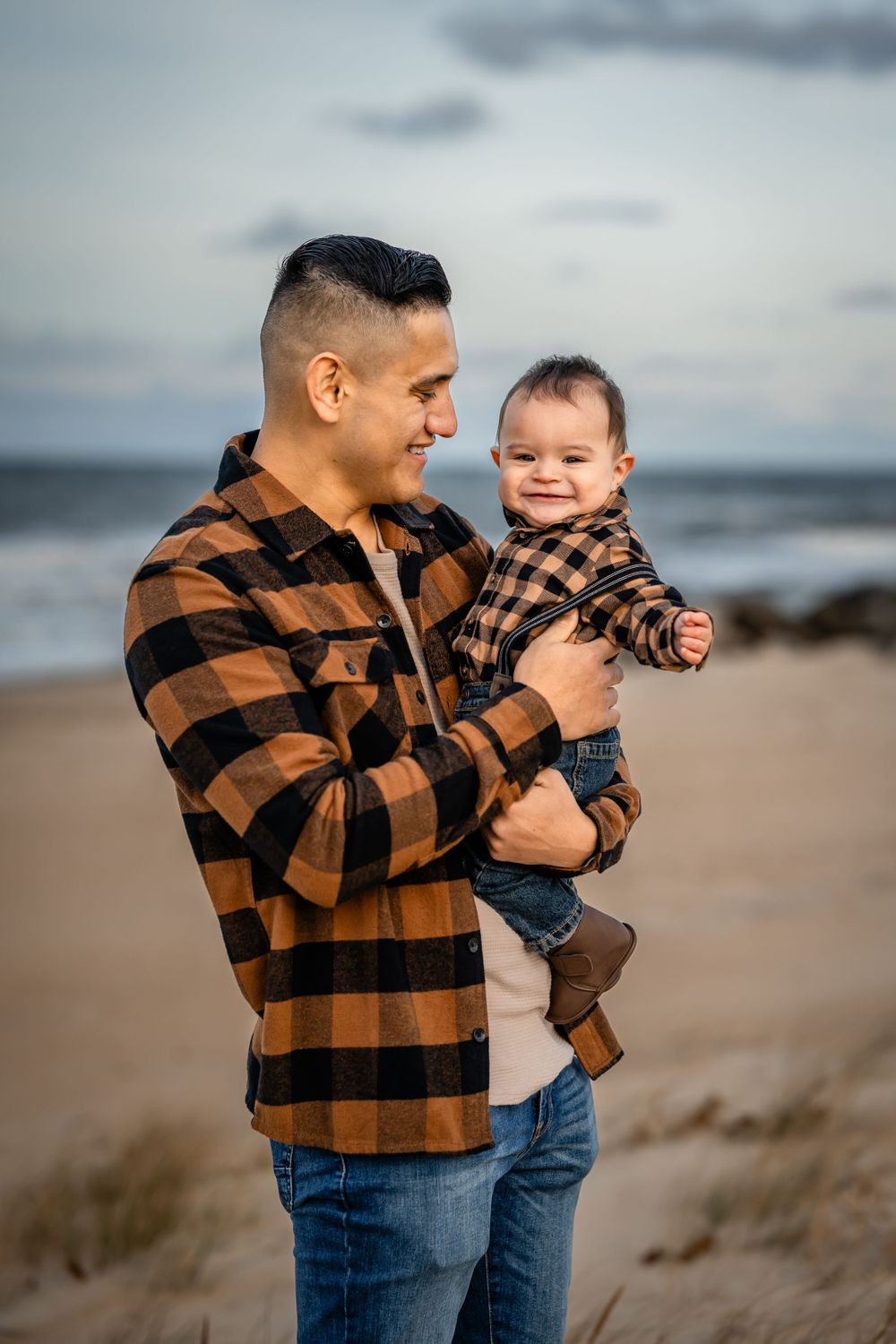 A tender moment between parent and baby wearing matching plaid shirts on a coastal beach.