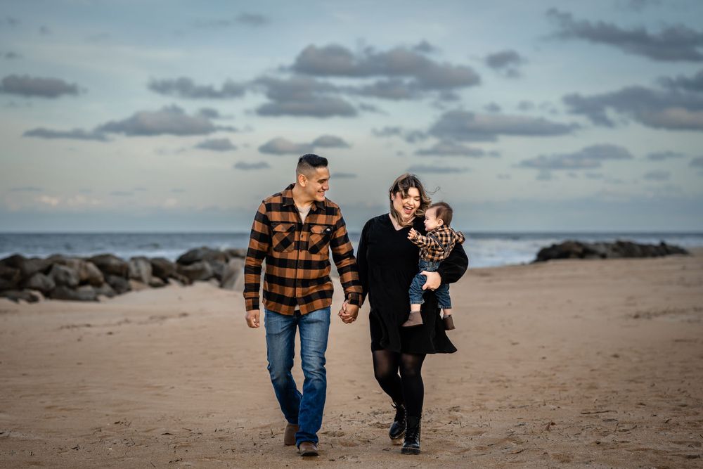A family walks together on a beach with rocky shoreline and cloudy sky in the background.