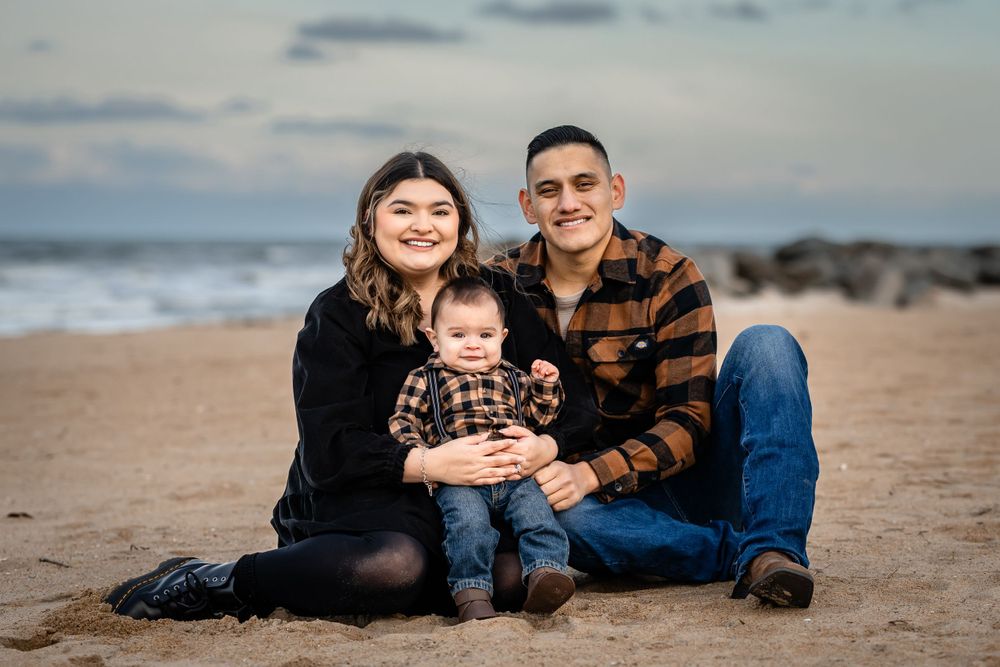 A family sits together on sandy beach sharing intimate moment with ocean and clouds in background.