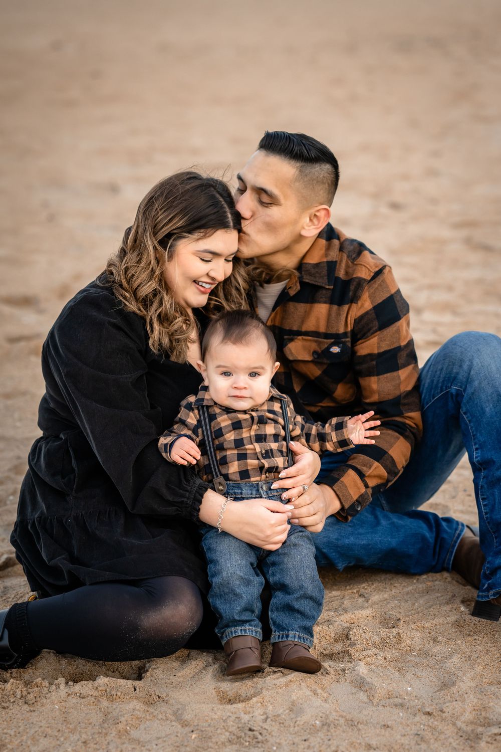 An intimate family portrait on beach sand with parents embracing their baby in matching plaid.