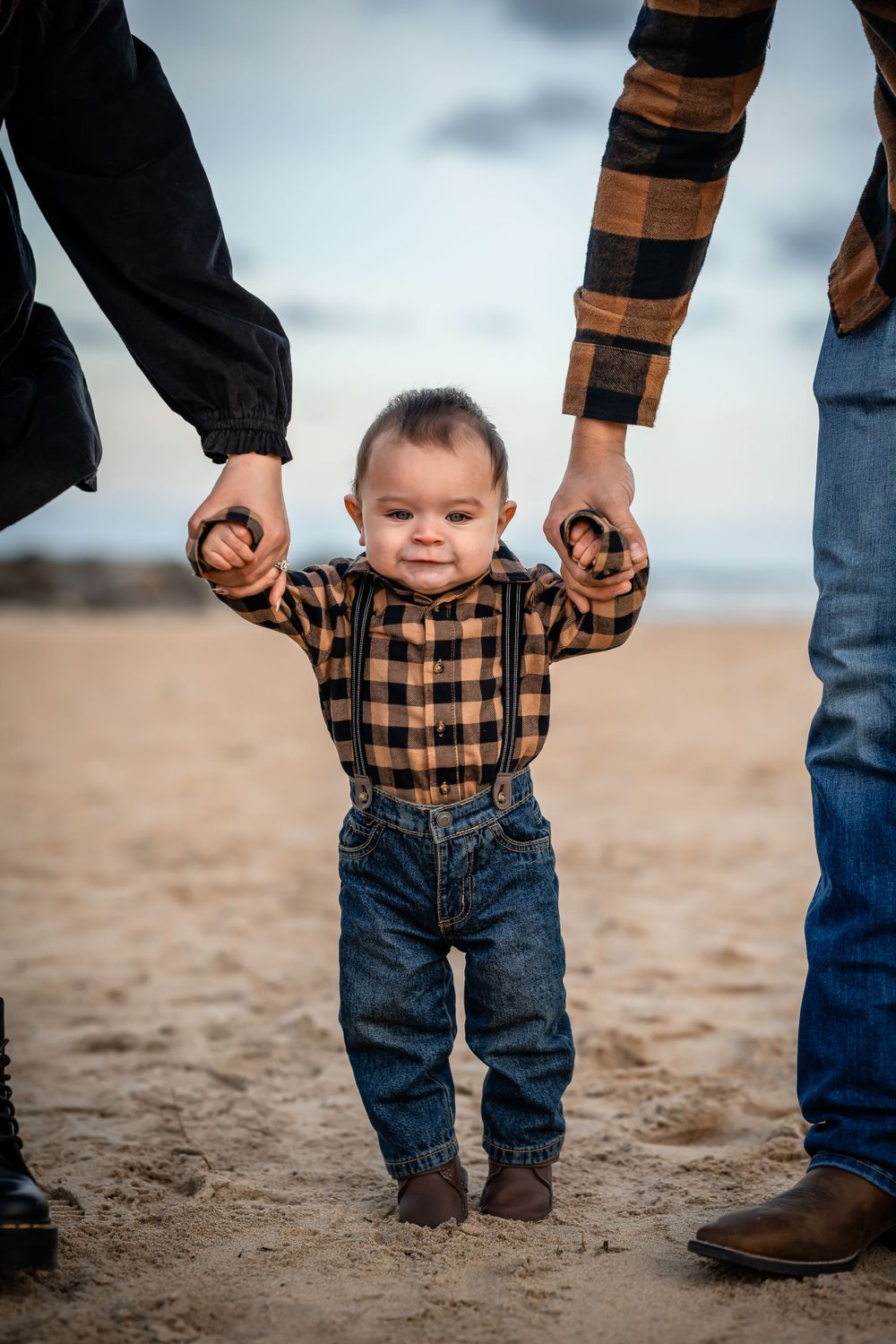 A small child in plaid shirt takes first steps holding parents' hands on sandy beach.