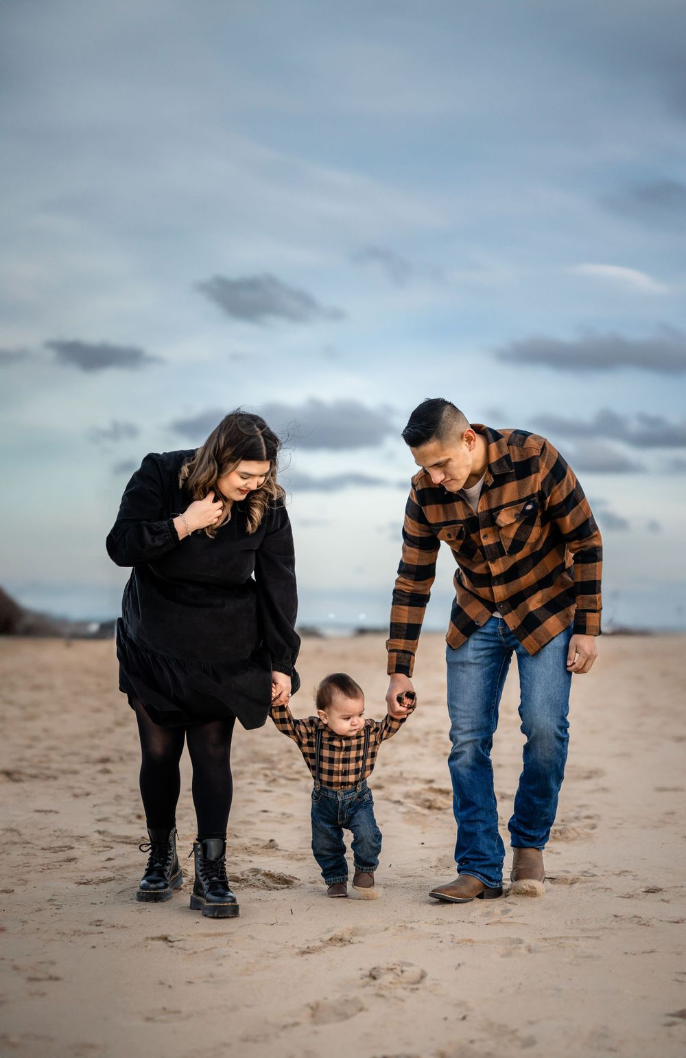 Parents help their toddler walk on beach sand while wearing coordinating plaid outfits.