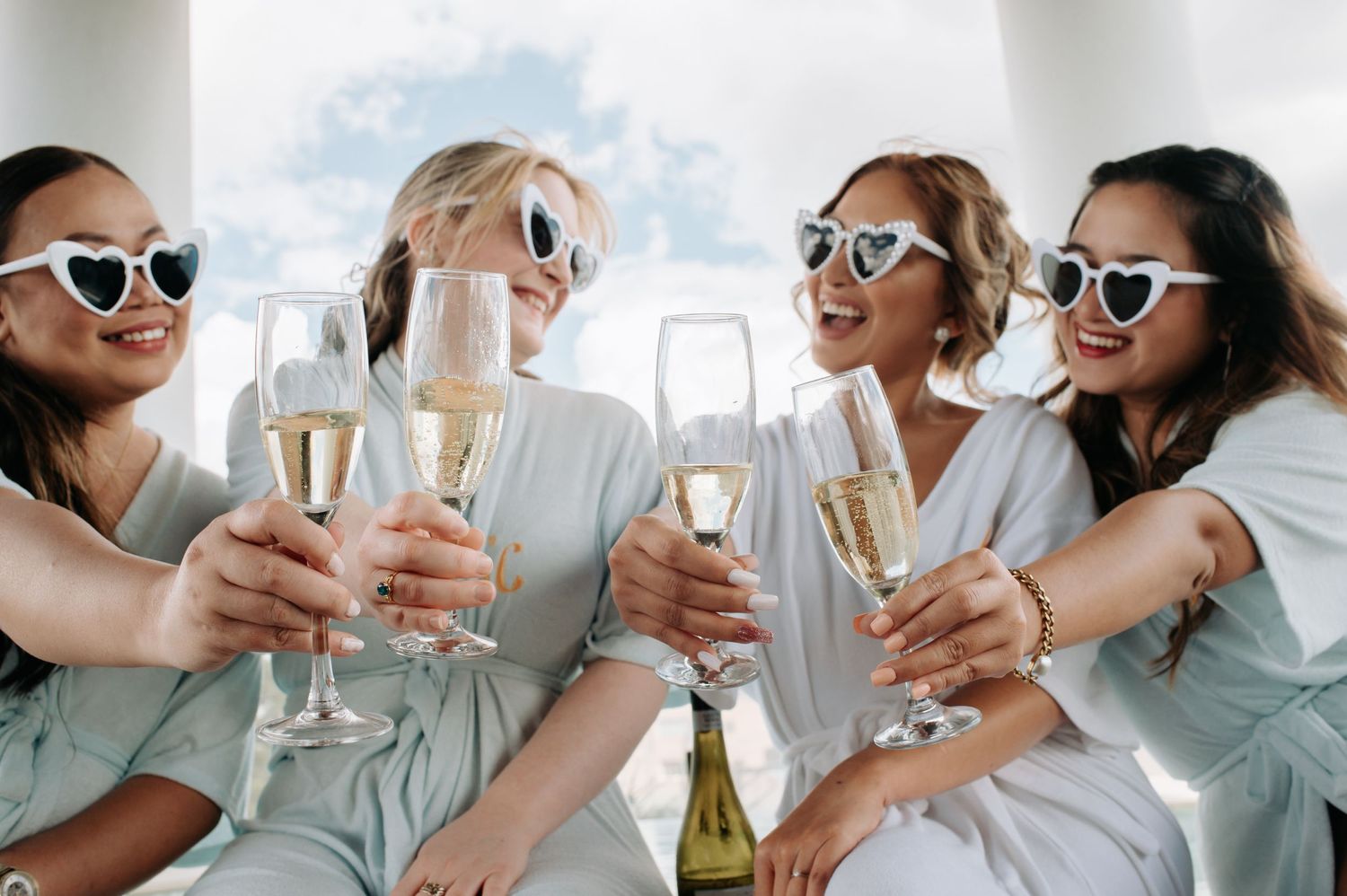 Friends in matching white outfits and sunglasses toasting with champagne glasses.