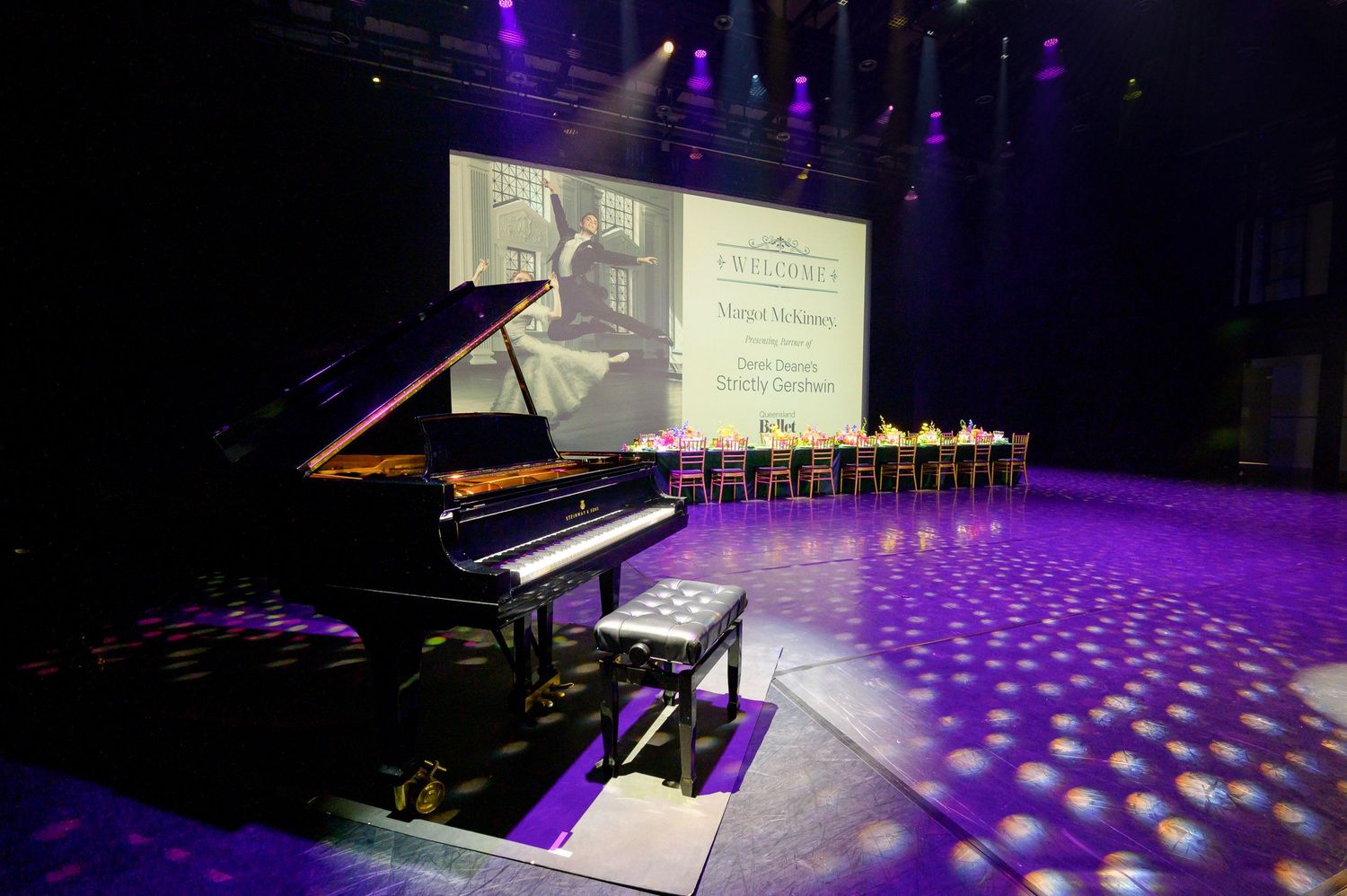 Grand piano on stage under purple lighting with projection screen and empty chairs in background.