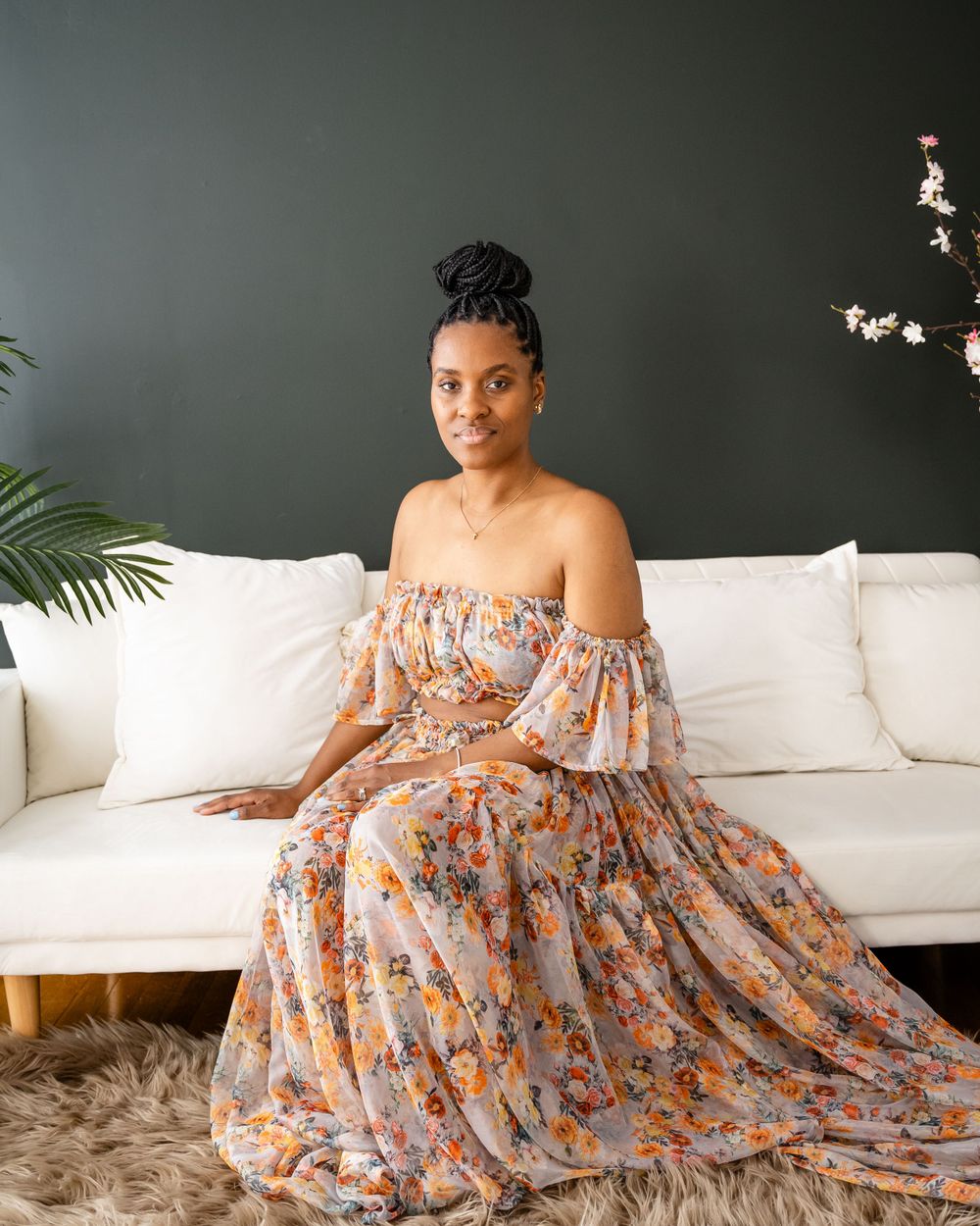 Elegant off-shoulder floral dress photographed on white sofa against dark grey wall.