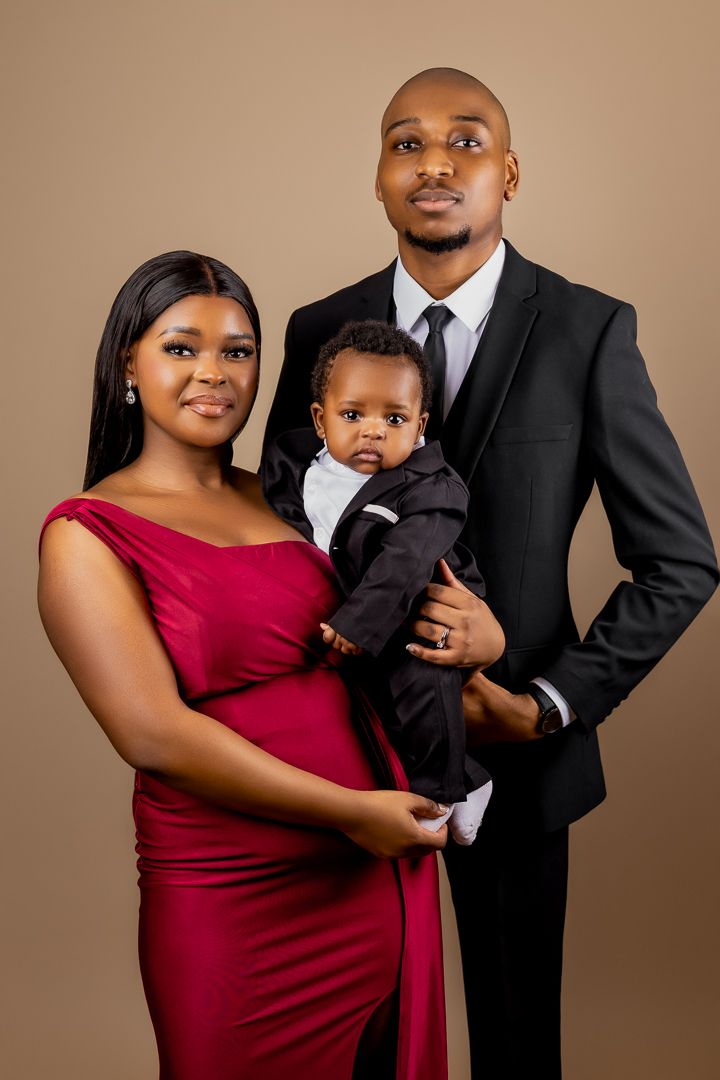 Elegant family portrait against brown backdrop featuring a red dress, black suit, and toddler in formal wear.