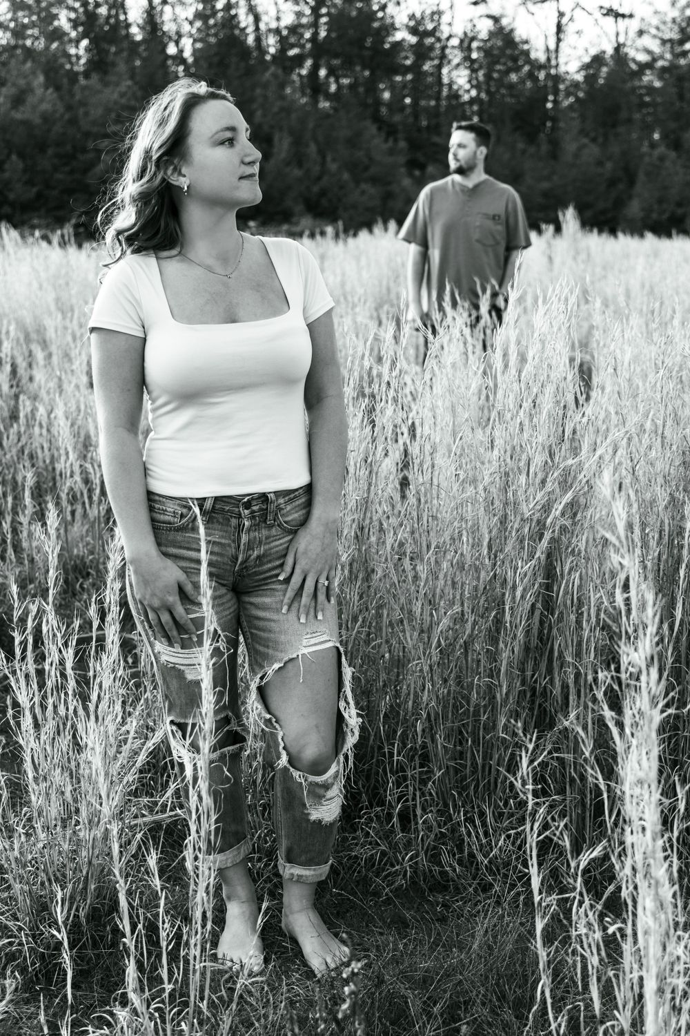 Artistic portrait of couple standing apart in rustic wheat field with one in the foreground and the other in the background.