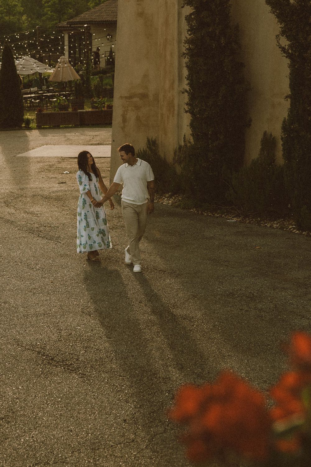 An engaged couple walking together on a paved path near pillars and red flowers in a romantic outdoor setting at a winery in vintage style.