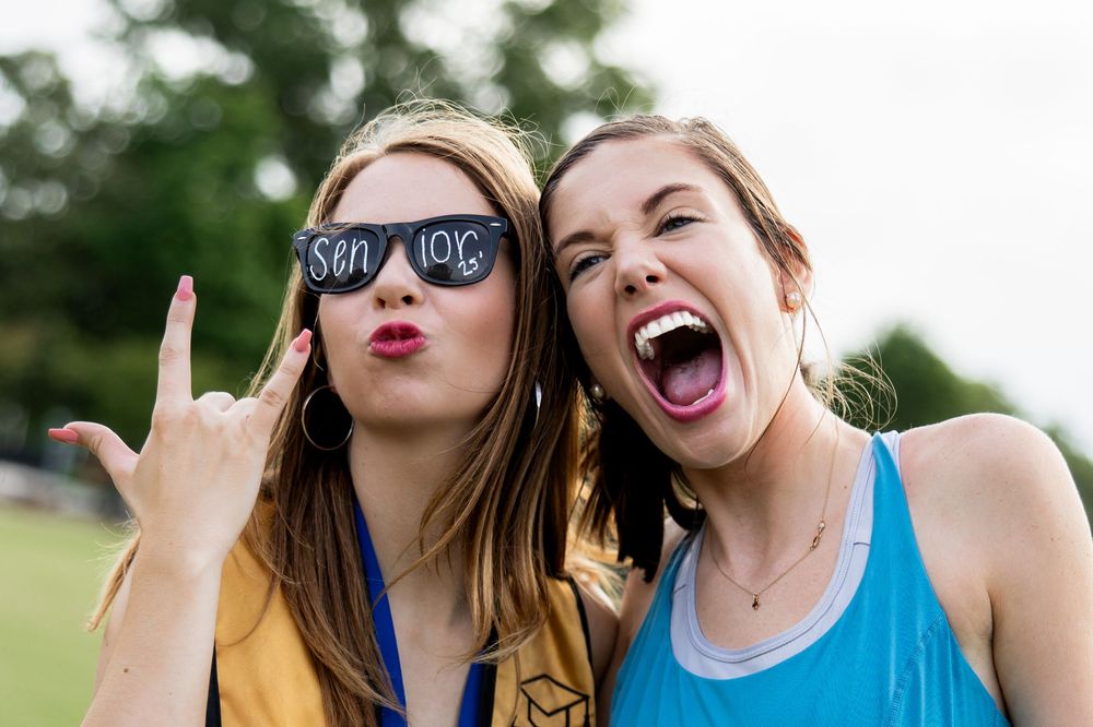 Two friends posing playfully outdoors during a senior grad session, one wearing sunglasses with text and making silly expressions.