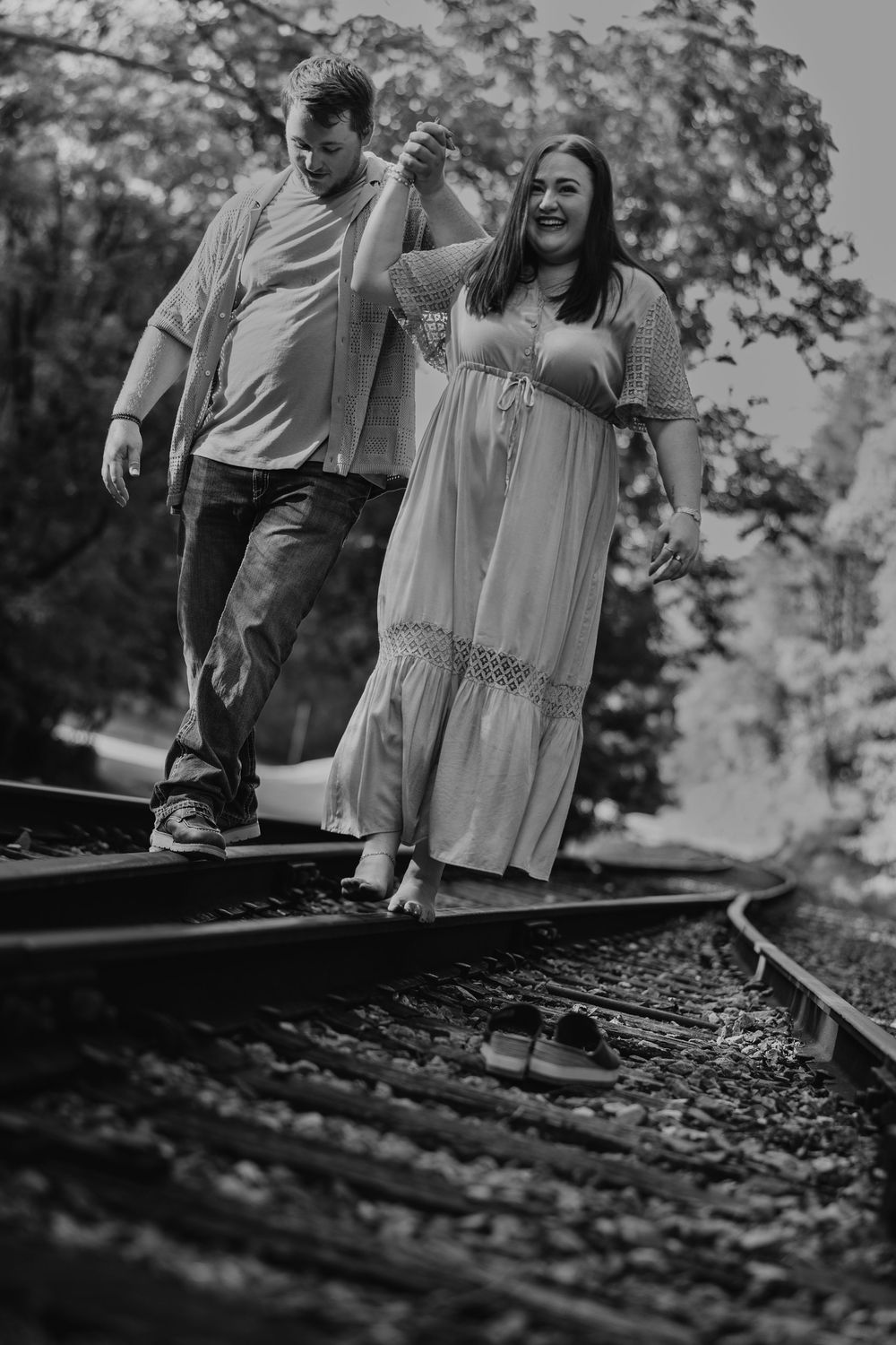 Black and white photo of couple walking and laughing together along train tracks in a romantic outdoor setting.