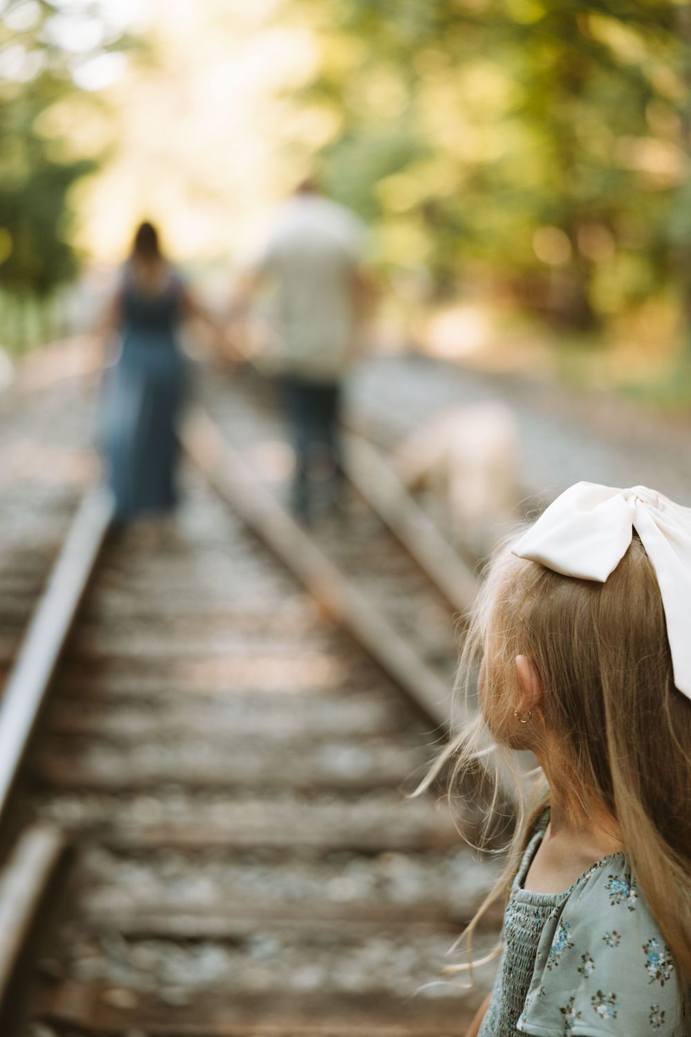A child with a white bow watches her parents walking on railway tracks in a dreamy summer setting.