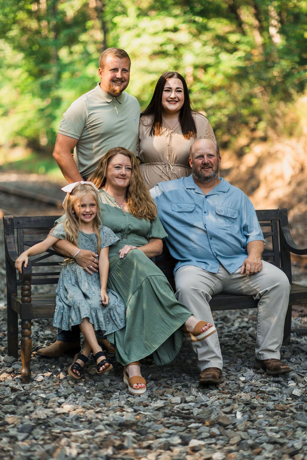 A family sits together on a rustic wooden bench in a sunlit forest setting pausing for candid moments during a photo shoot.